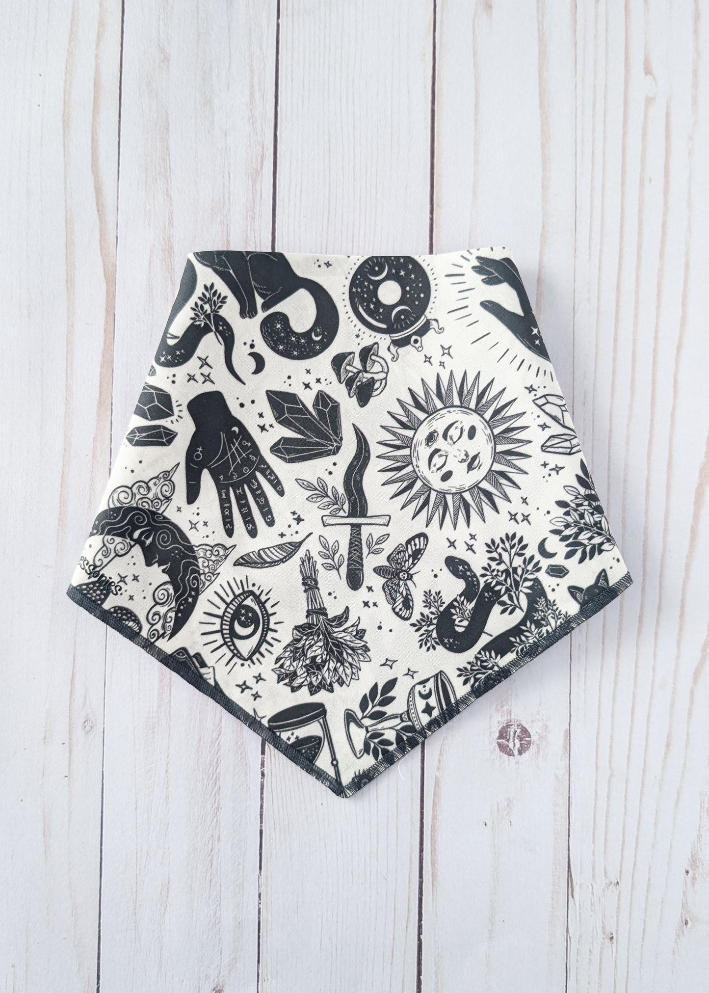 A triangular bandana-style bib or scarf with a black and white witchy-themed print, laid flat on a light whitewashed wooden background.