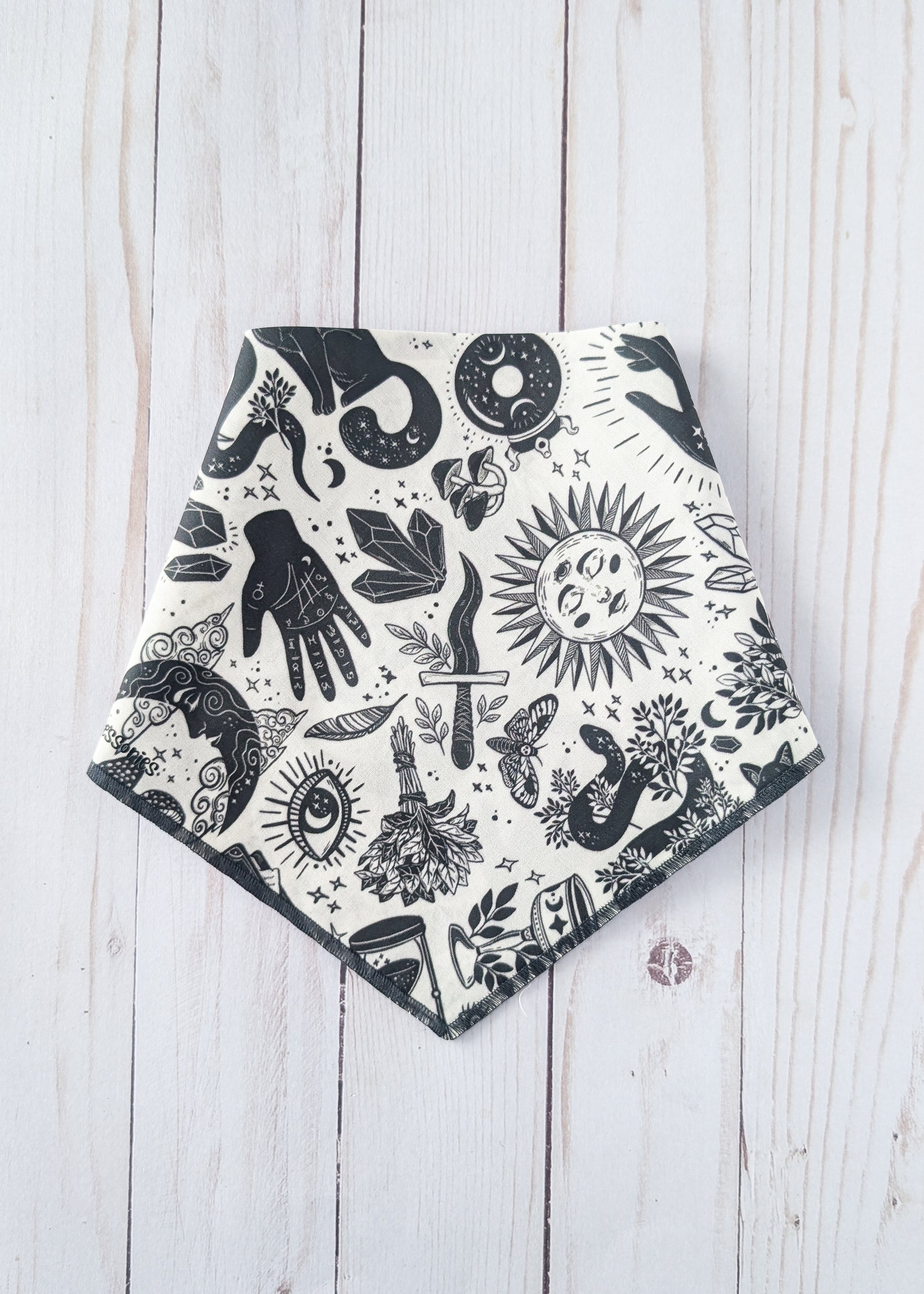 A triangular bandana-style bib or scarf with a black and white witchy-themed print, laid flat on a light whitewashed wooden background.