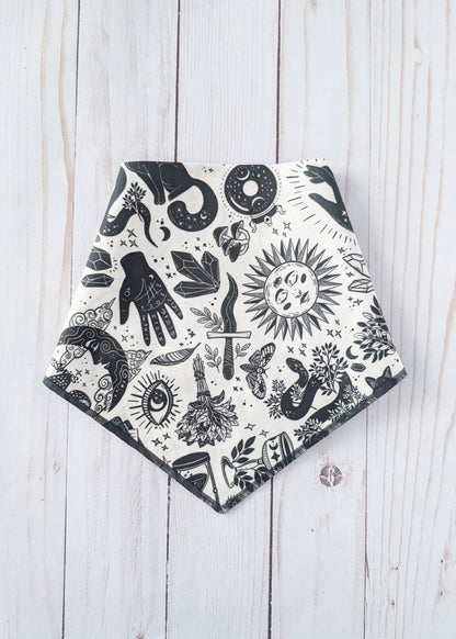 A triangular bandana-style bib or scarf with a black and white witchy-themed print, laid flat on a light whitewashed wooden background.