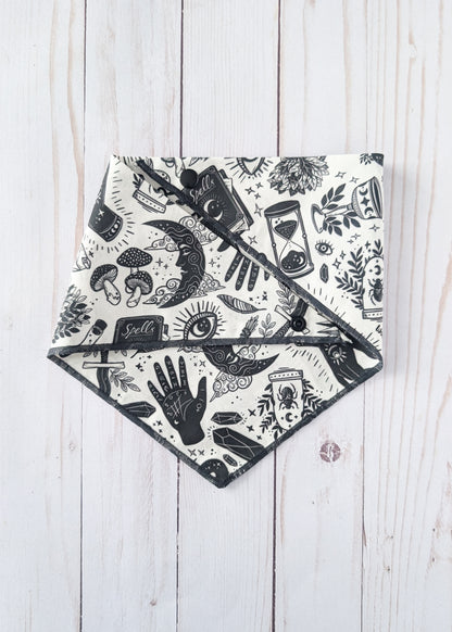 A triangular bandana-style bib or scarf with a black and white witchy-themed print, laid flat on a light whitewashed wooden background.