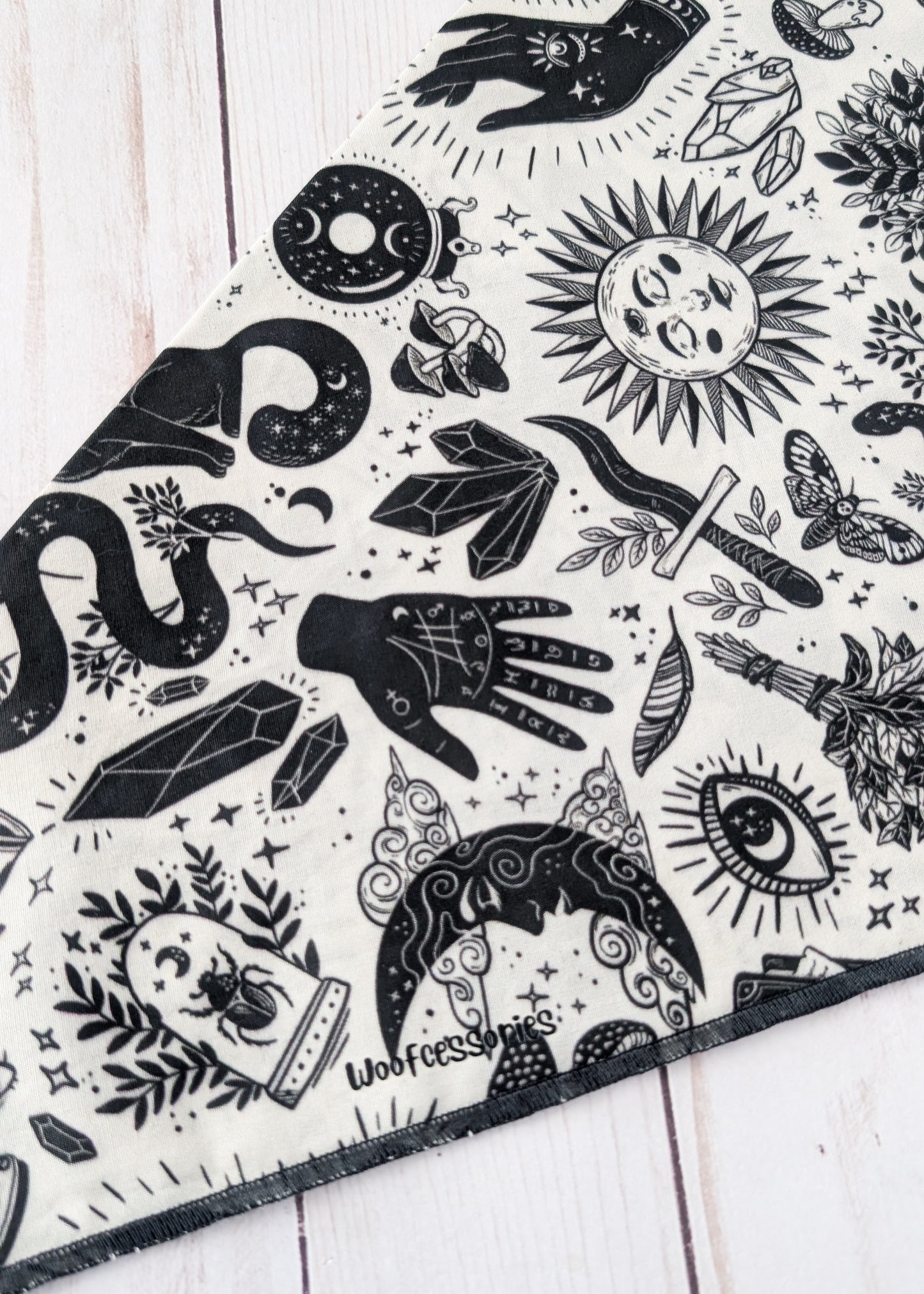 A close-up of a black and white patterned bandana or scarf laid diagonally on a white wooden surface. The fabric features a detailed graphic print with wtichy and mystical illustrations including a snake, crystal, dagger, eye, moon, and hand