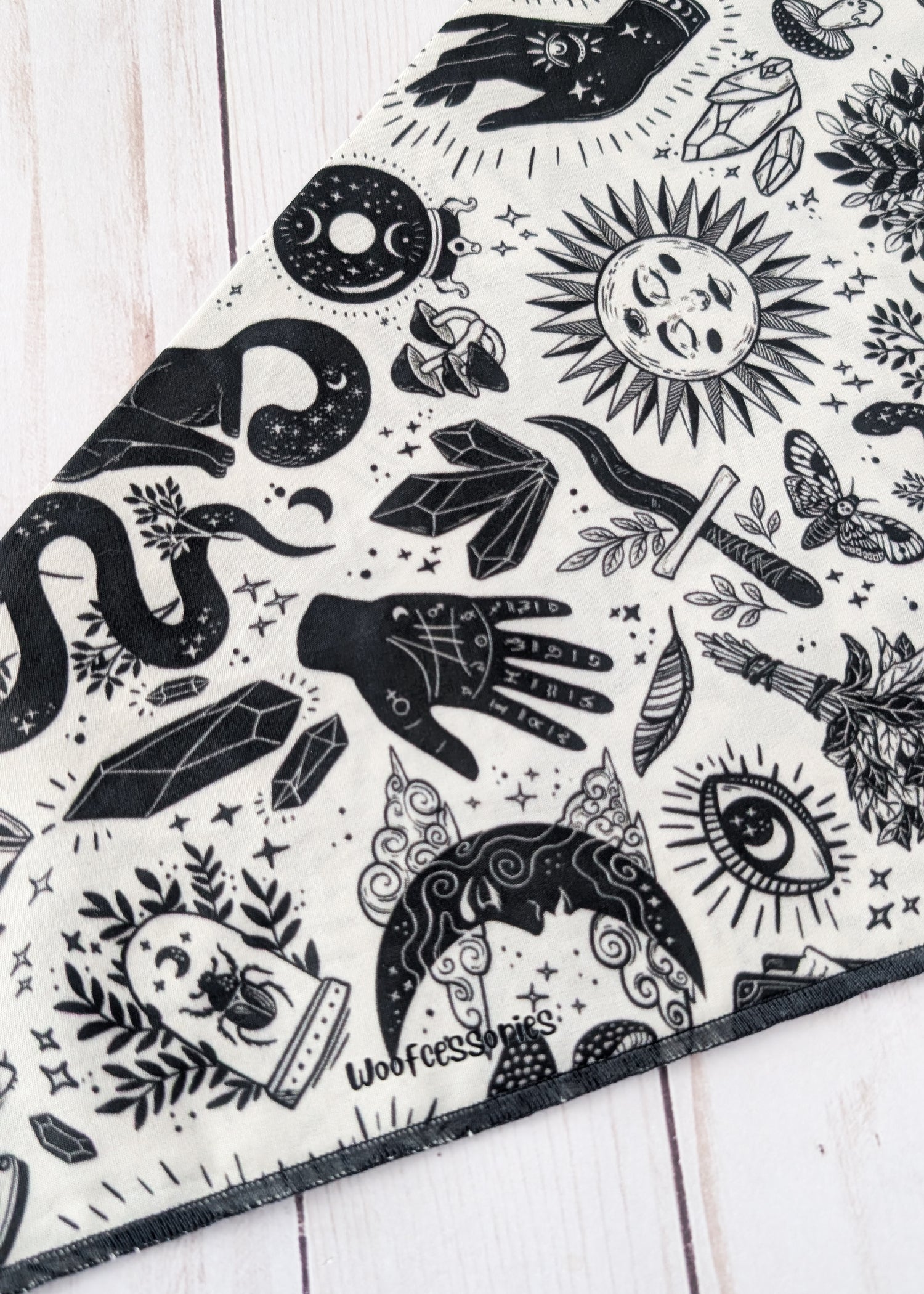 A close-up of a black and white patterned bandana or scarf laid diagonally on a white wooden surface. The fabric features a detailed graphic print with wtichy and mystical illustrations including a snake, crystal, dagger, eye, moon, and hand