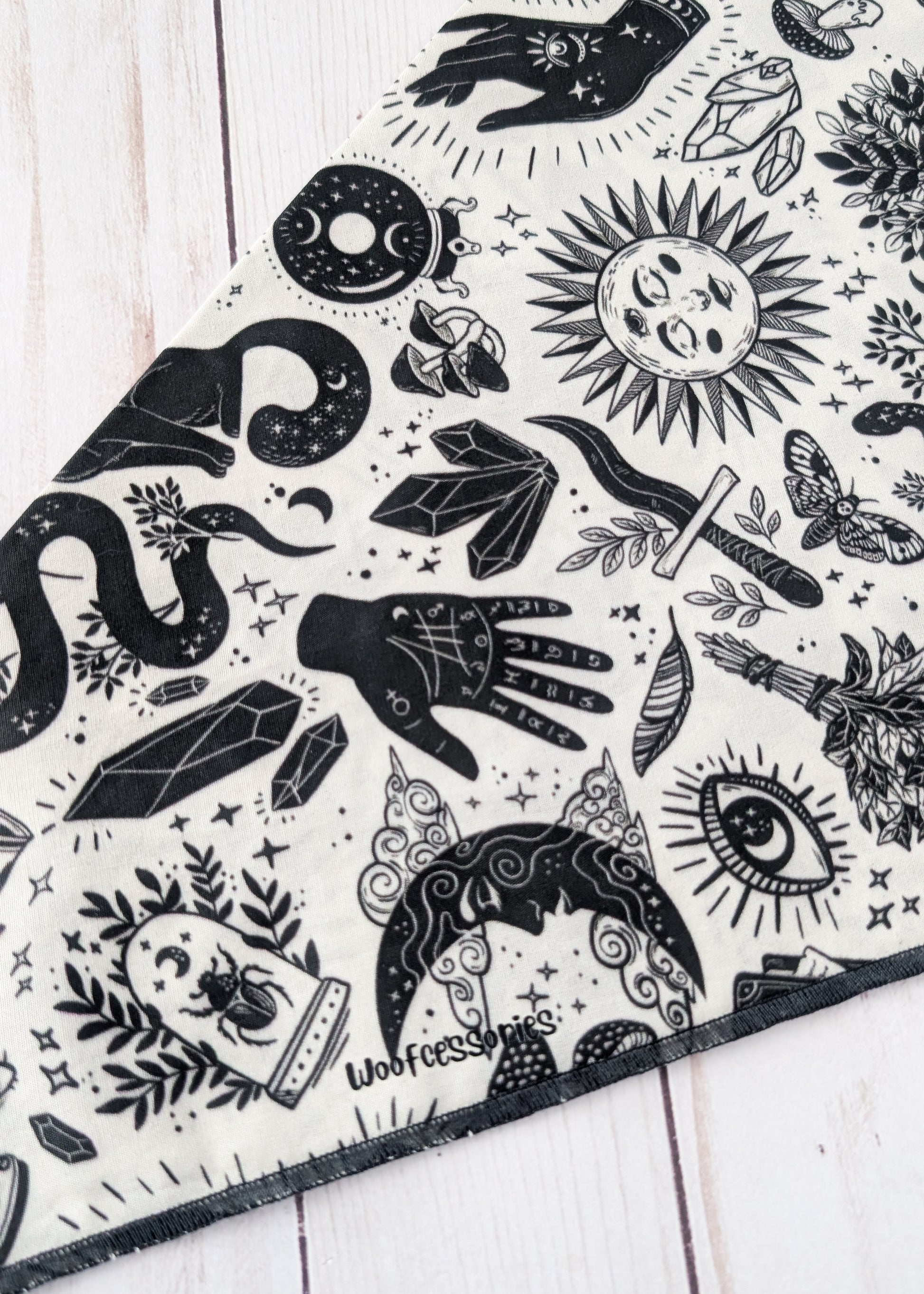 A close-up of a black and white patterned bandana or scarf laid diagonally on a white wooden surface. The fabric features a detailed graphic print with wtichy and mystical illustrations including a snake, crystal, dagger, eye, moon, and hand
