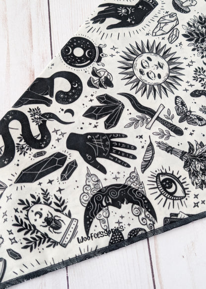 A close-up of a black and white patterned bandana or scarf laid diagonally on a white wooden surface. The fabric features a detailed graphic print with wtichy and mystical illustrations including a snake, crystal, dagger, eye, moon, and hand