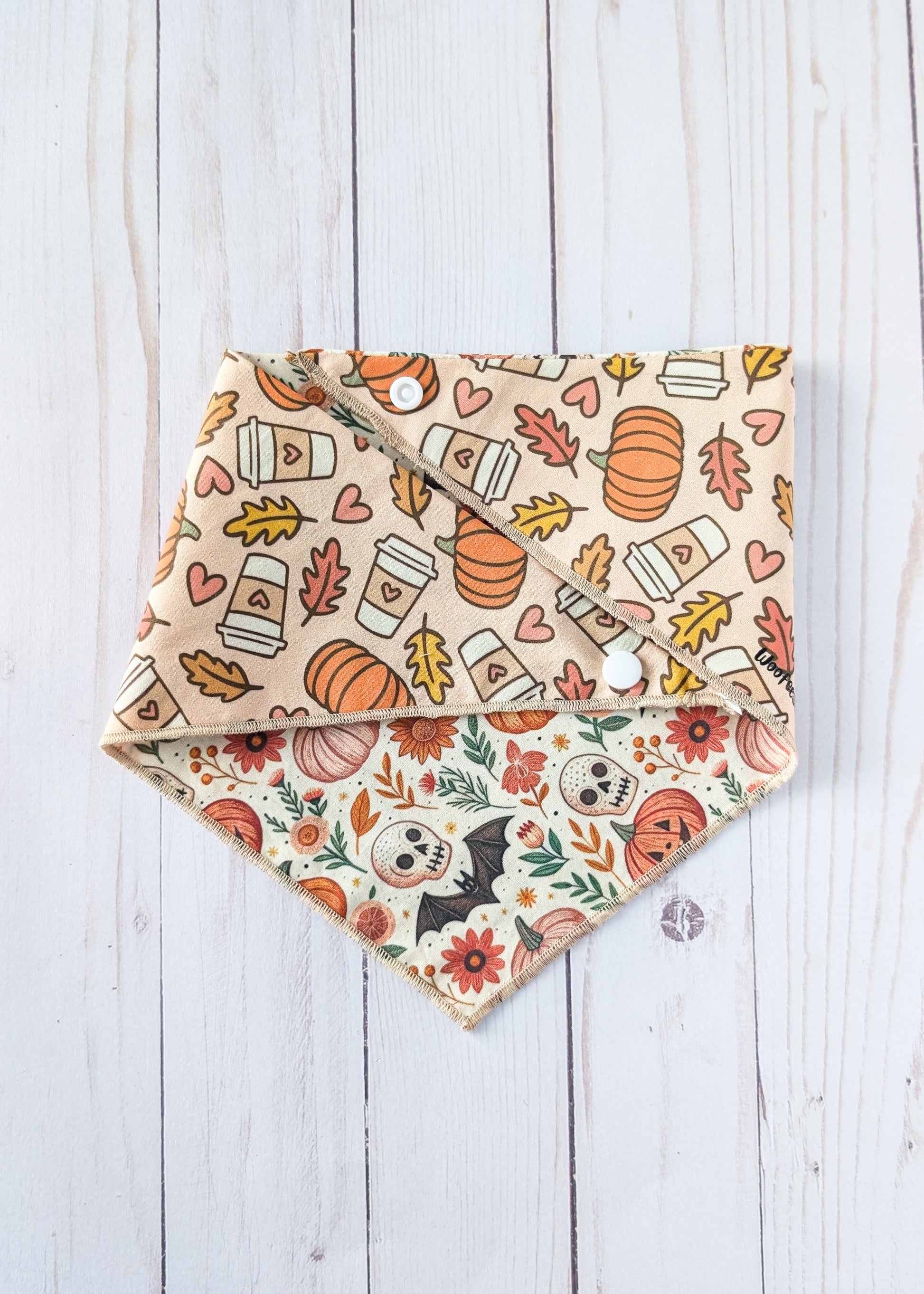a triangle bandanas with autumn Halloween patterns, displayed on a white wood plank background. One bandana features orange pumpkins, coffee cups, and hearts on a beige background, while the other features orange pumpkins, skulls, flowers, and bats on a white background.