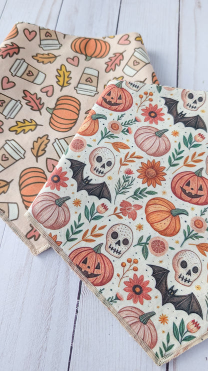 Two bandanas  on a white wooden surface. One bandana features a light beige background with orange pumpkins, coffee cups, and autumn leaves.