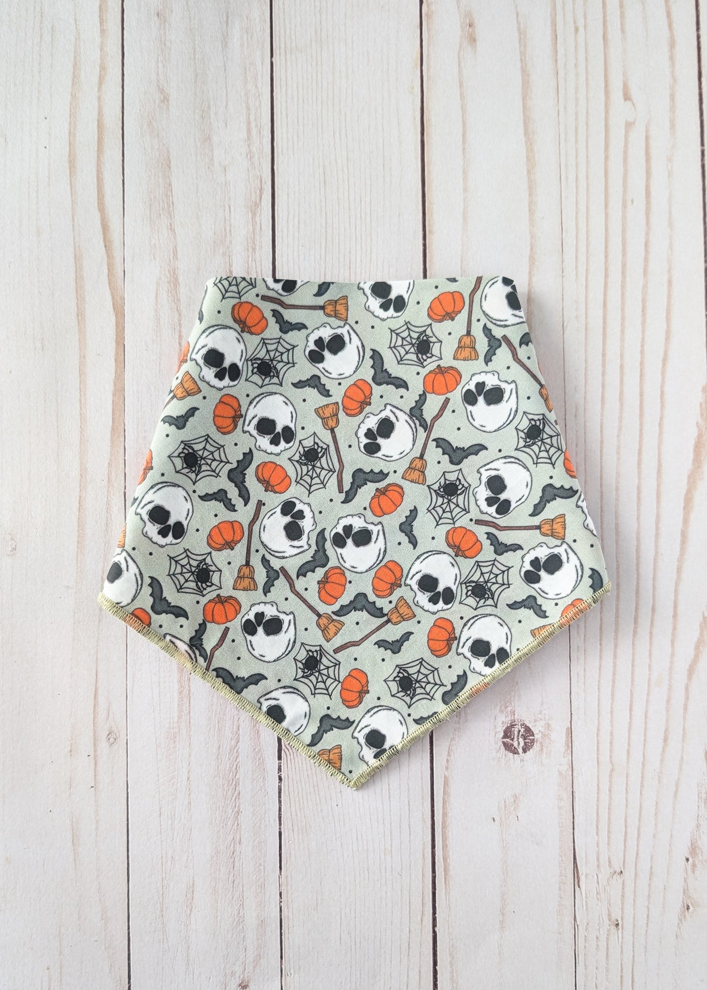 A triangular bandana bib with a Halloween pattern of skulls, pumpkins, spiderwebs, and bats on a sage green background, laid flat on a light wood plank surface.