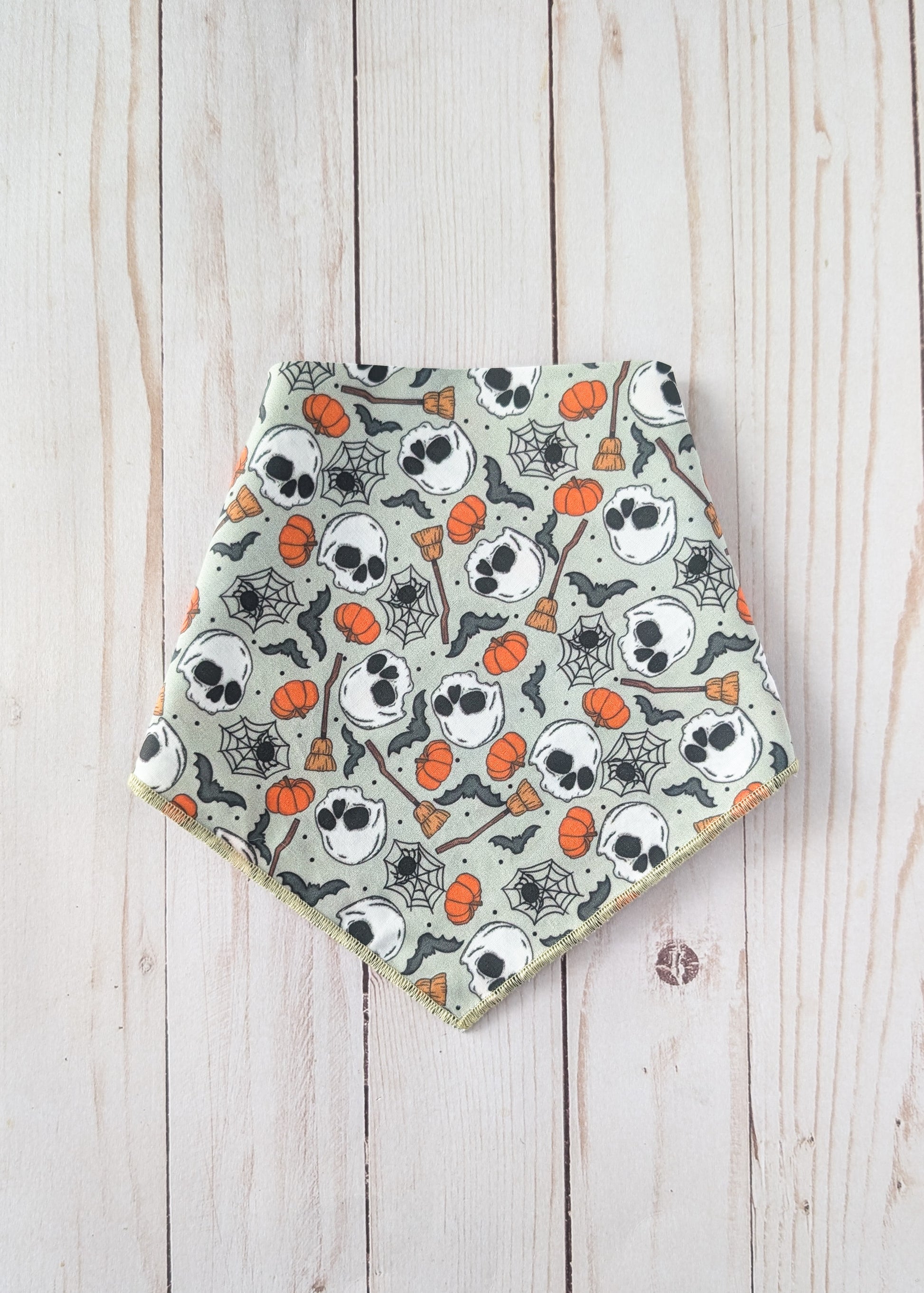 A triangular bandana bib with a Halloween pattern of skulls, pumpkins, spiderwebs, and bats on a sage green background, laid flat on a light wood plank surface.