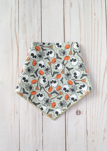 A triangular bandana bib with a Halloween pattern of skulls, pumpkins, spiderwebs, and bats on a sage green background, laid flat on a light wood plank surface.