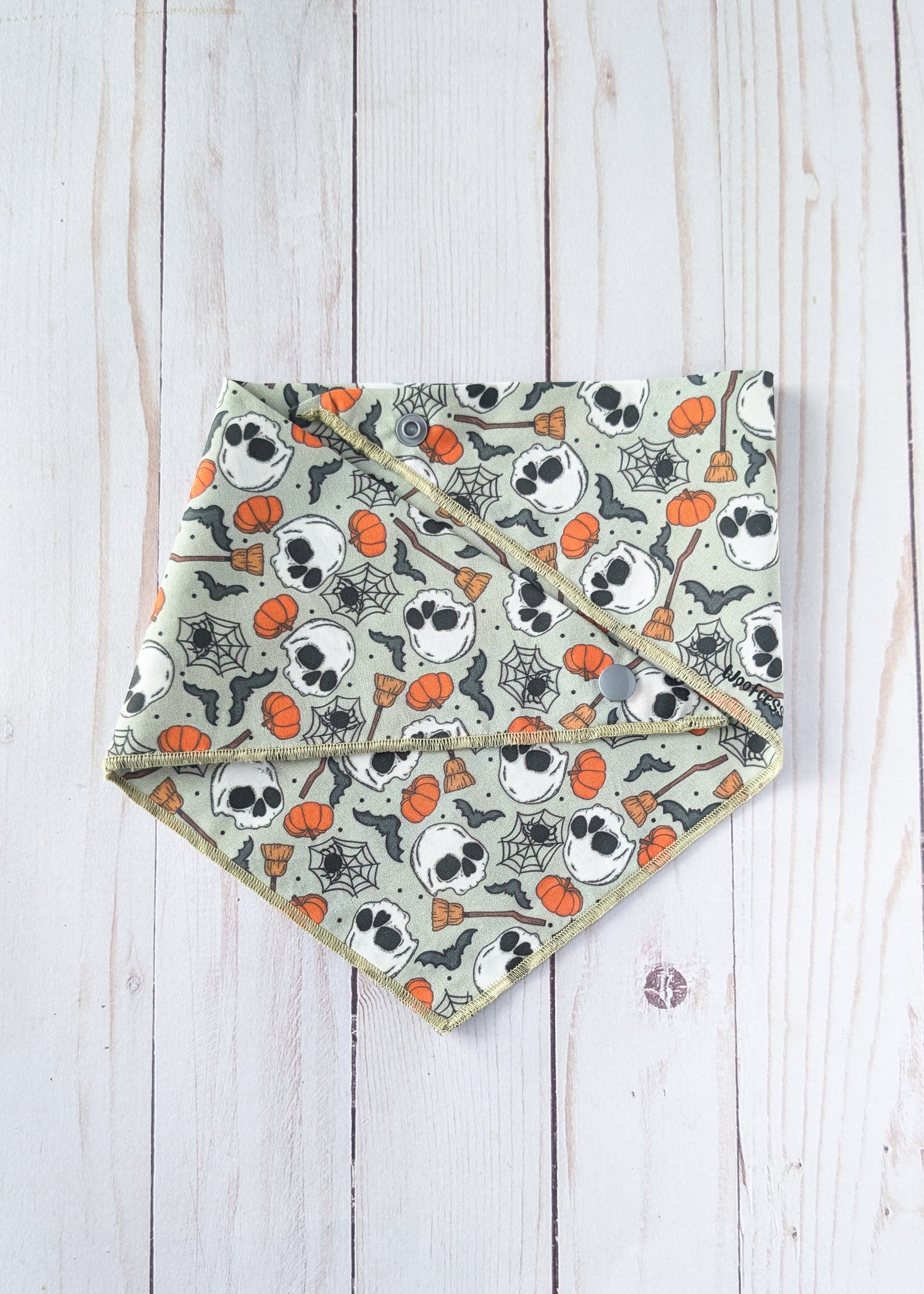 A triangular bandana-style pet bib with a Halloween print of skulls, pumpkins, bats, and spiders on a light sage green background, laid flat on a white wood plank surface.