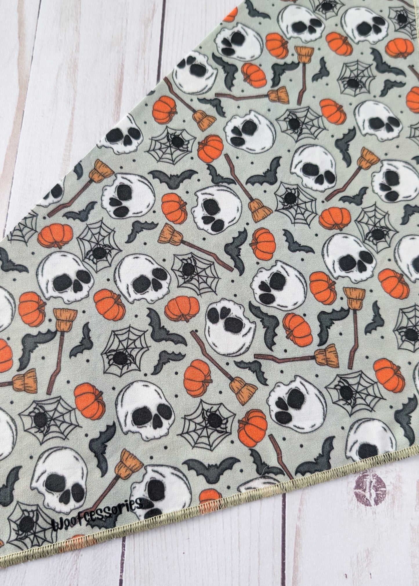 A triangular bandana with a Halloween pattern of skulls, pumpkins, spiderwebs, and brooms on a sage green background, laid flat on a white wood plank surface.