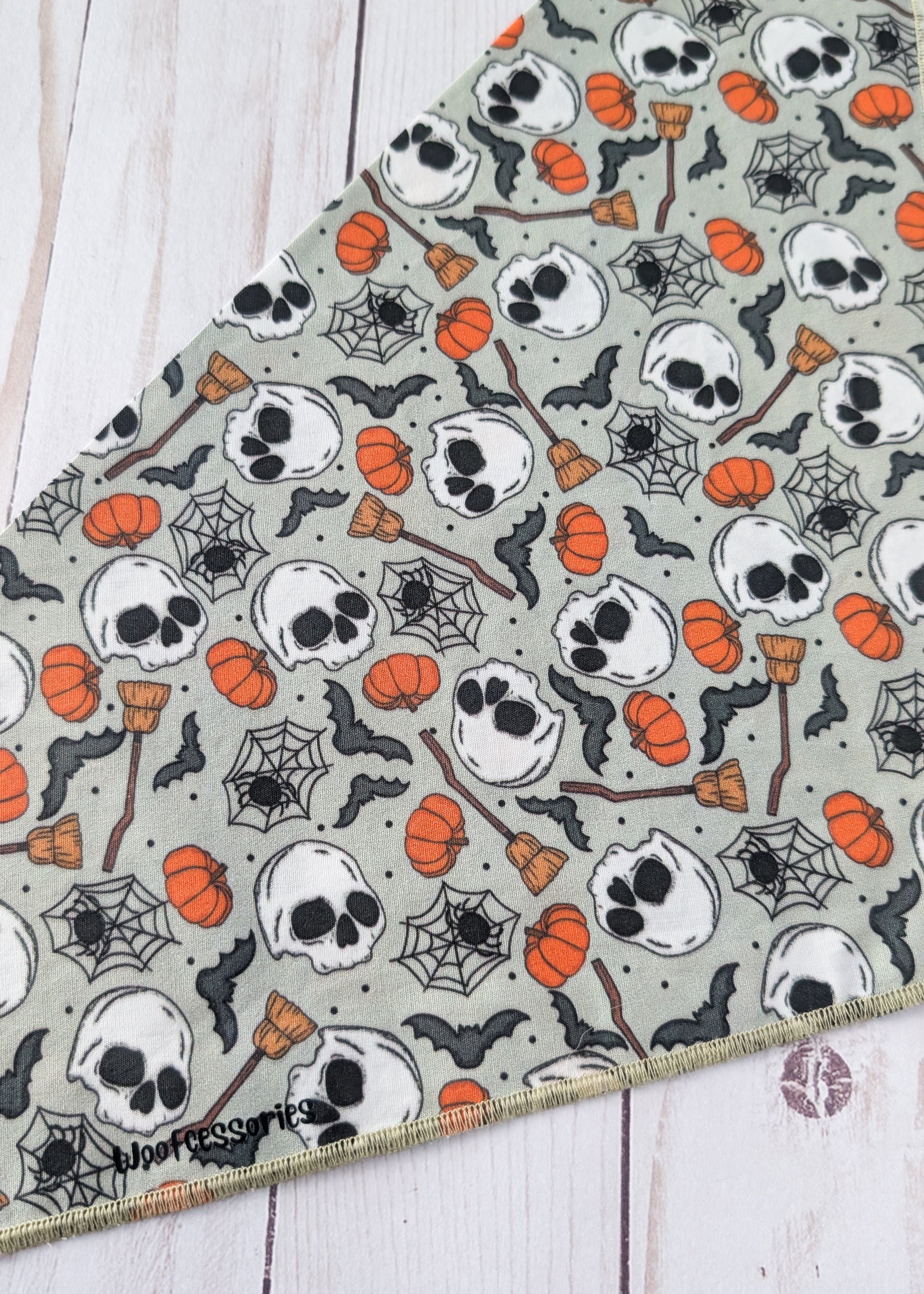 A triangular bandana with a Halloween pattern of skulls, pumpkins, spiderwebs, and brooms on a sage green background, laid flat on a white wood plank surface.