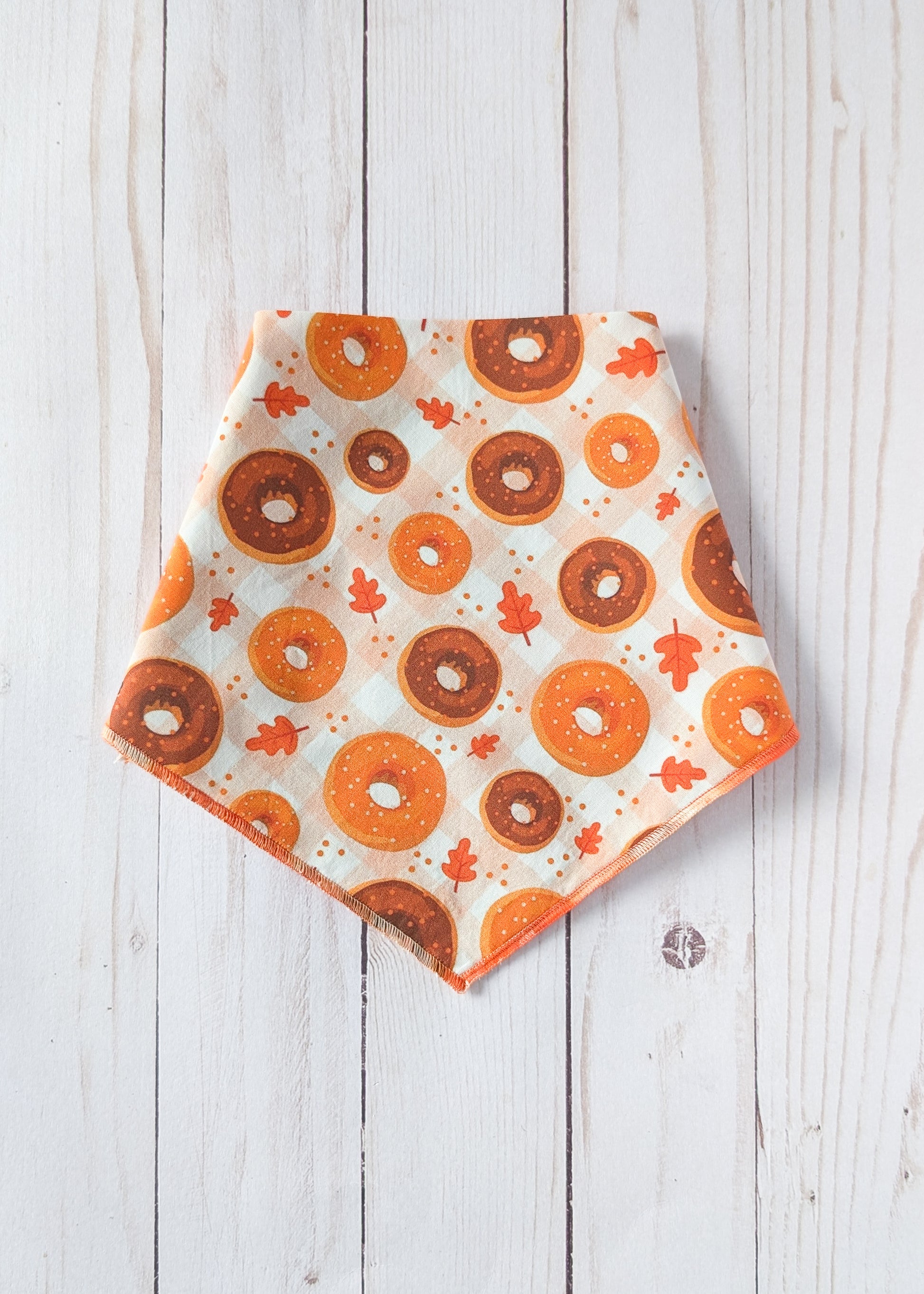 A triangular bandana with an orange and brown donut pattern and small orange leaves on an off-white background, laid flat on a white wood plank surface.