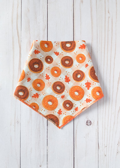 A triangular bandana with an orange and brown donut pattern and small orange leaves on an off-white background, laid flat on a white wood plank surface.