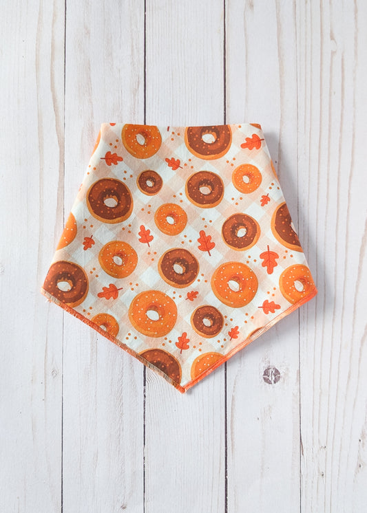 A triangular bandana with an orange and brown donut pattern and small orange leaves on an off-white background, laid flat on a white wood plank surface.