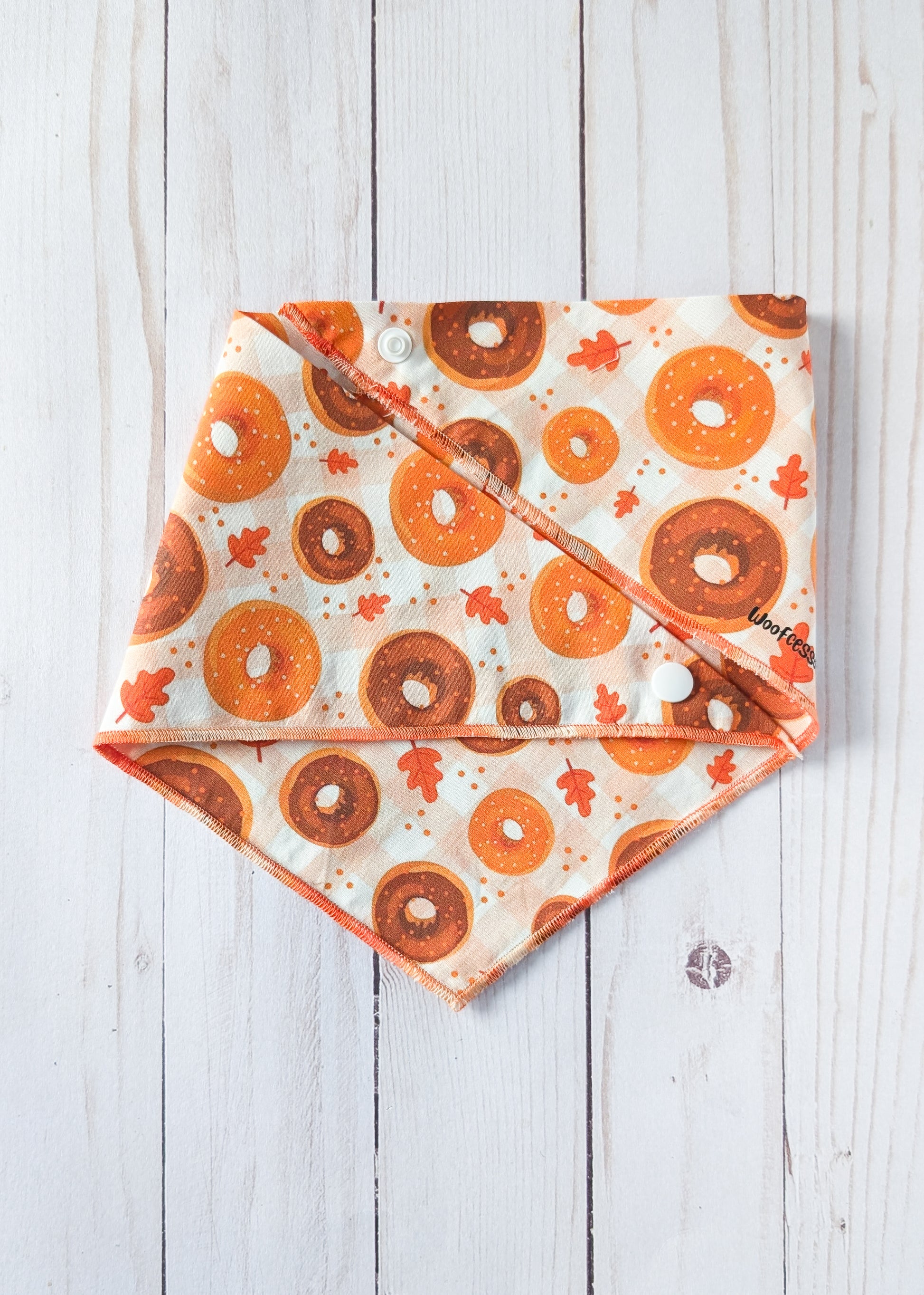 A triangular pet bandana with a printed pattern of orange and brown donuts and small orange leaves on an off-white background, laid flat on a white wood plank surface.