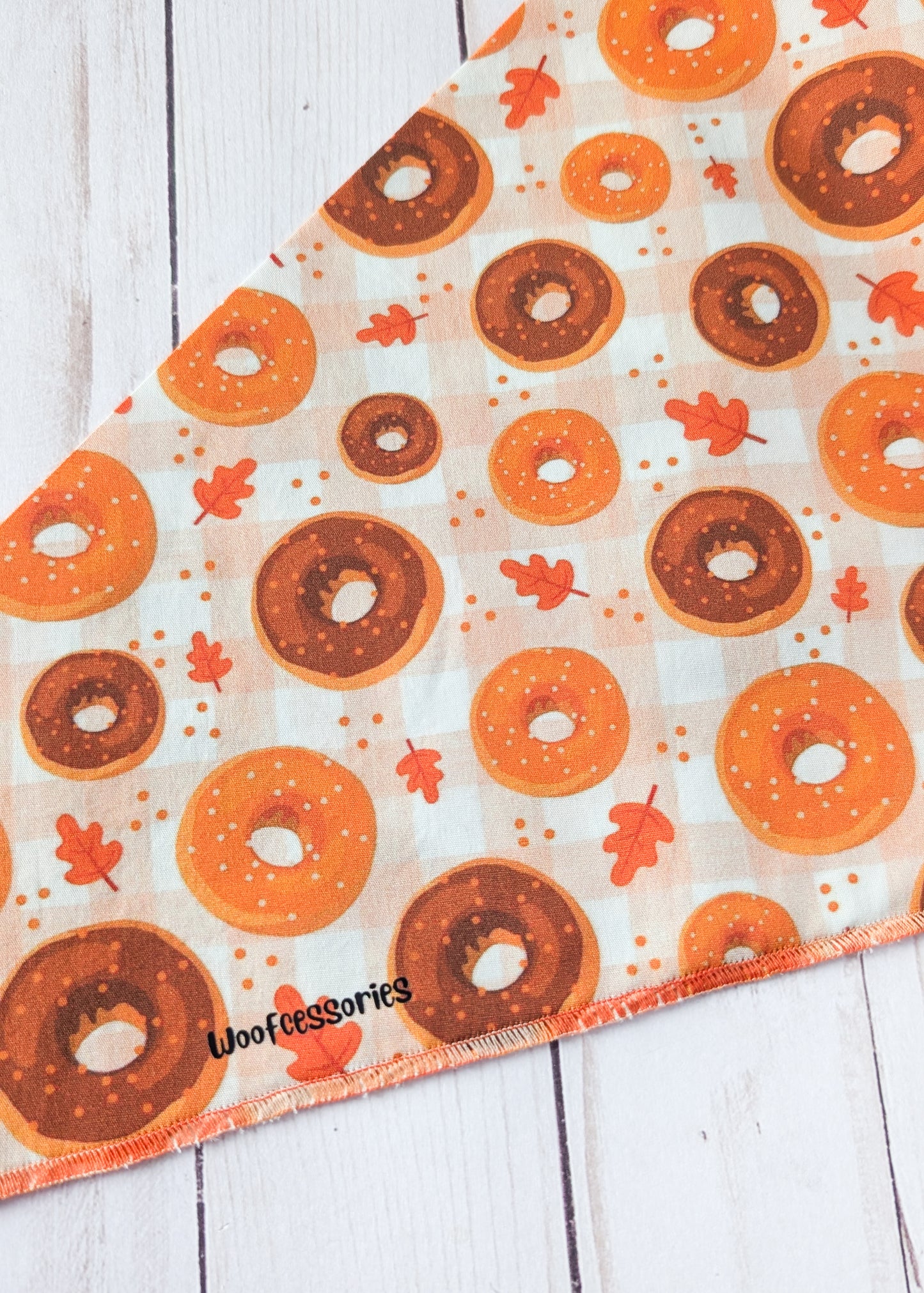 A triangular pet bandana close up with a donut and autumn leaf pattern, displayed on a white wood plank background. The bandana features orange, brown, and white donuts with small orange maple leaves and dots on a white background.
