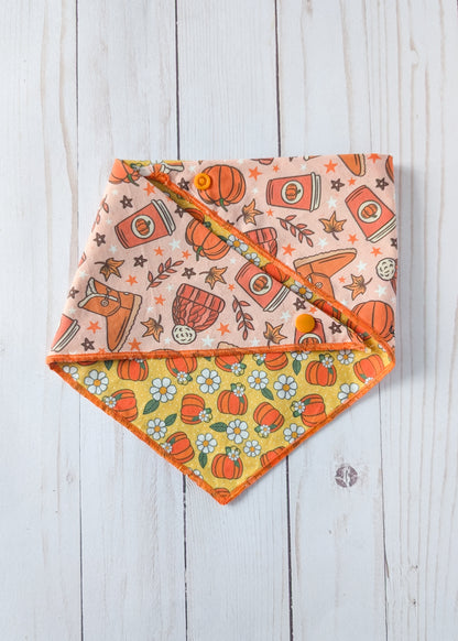 A triangular bandana-style pet bib laid flat on a white wood plank background, showing two printed sides: one in peach with orange pumpkins, stars, leaves, and a coffee cup, and one in yellow with the same pumpkin and white flower pattern.