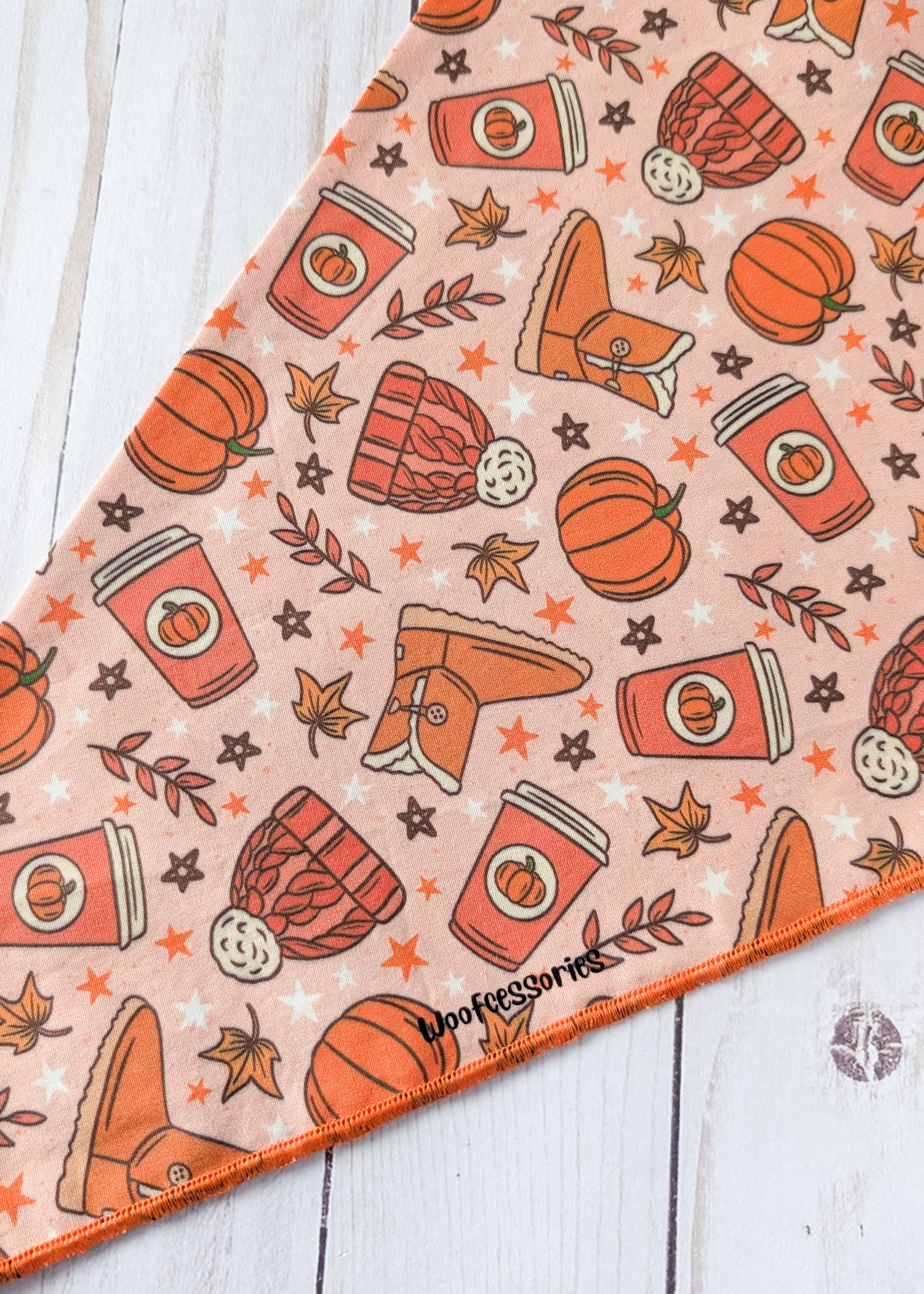 A triangular bandana with an autumn-themed pattern of pumpkins, coffee cups, boots, stars, and leaves on a peach background, laid flat on a white wood surface.