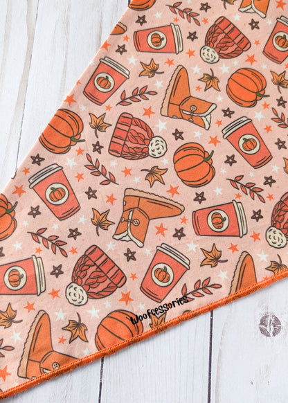 A triangular bandana with an autumn-themed pattern of pumpkins, coffee cups, boots, stars, and leaves on a peach background, laid flat on a white wood surface.