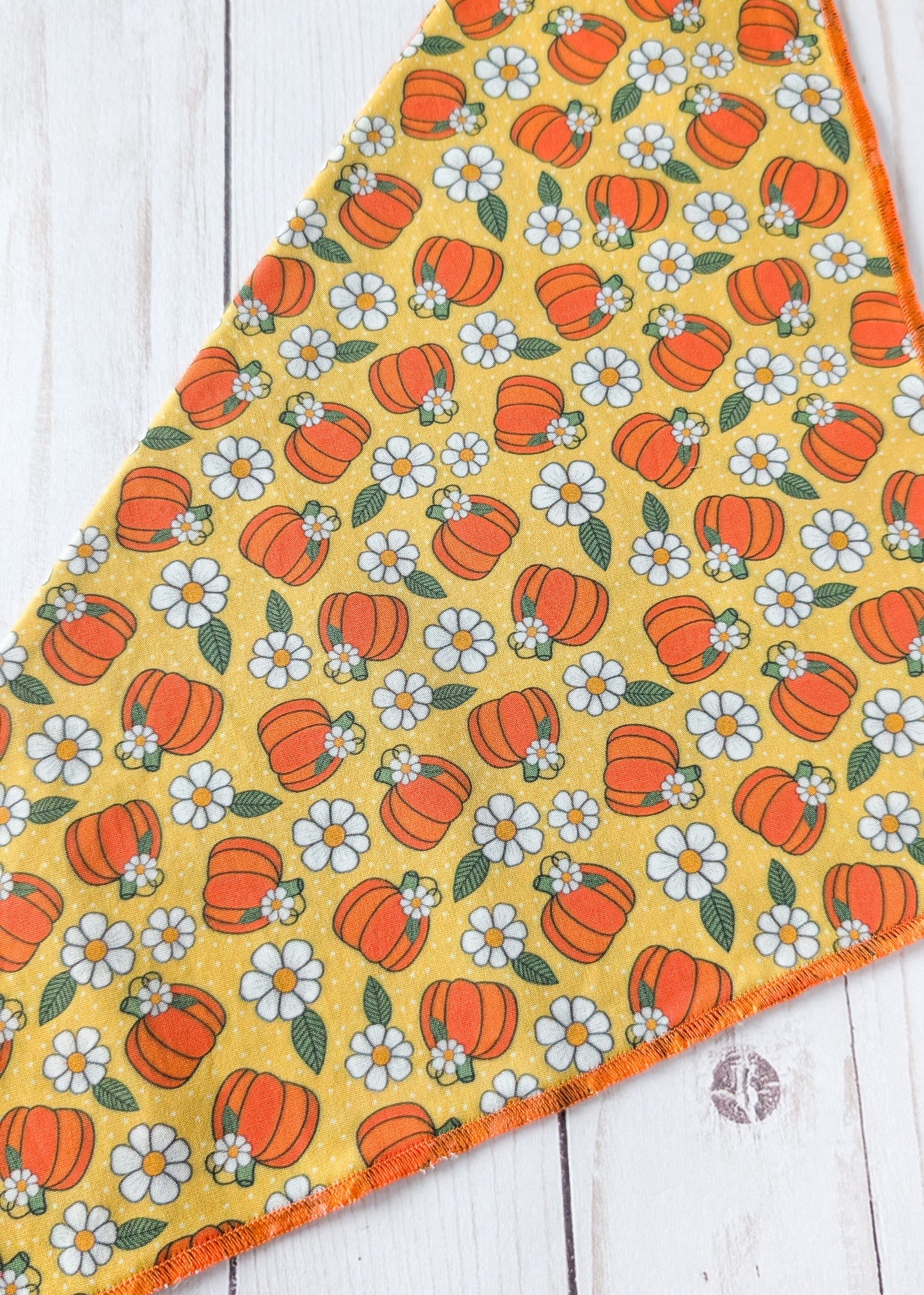 A triangular bandana, featuring a repeating pattern of orange pumpkins and white daisies with green leaves on a mustard yellow background, laid flat on a white wood plank surface.