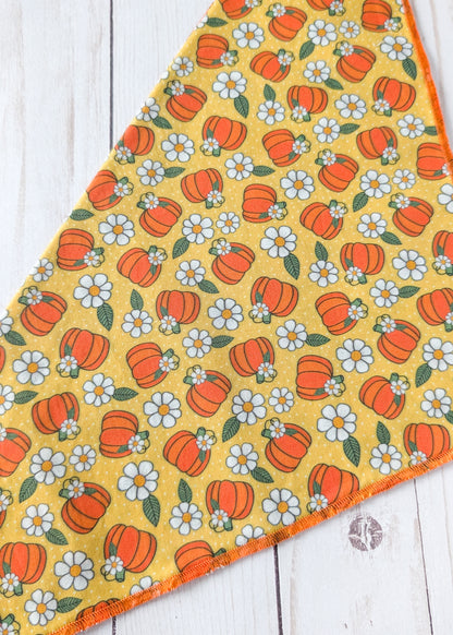 A triangular bandana, featuring a repeating pattern of orange pumpkins and white daisies with green leaves on a mustard yellow background, laid flat on a white wood plank surface.