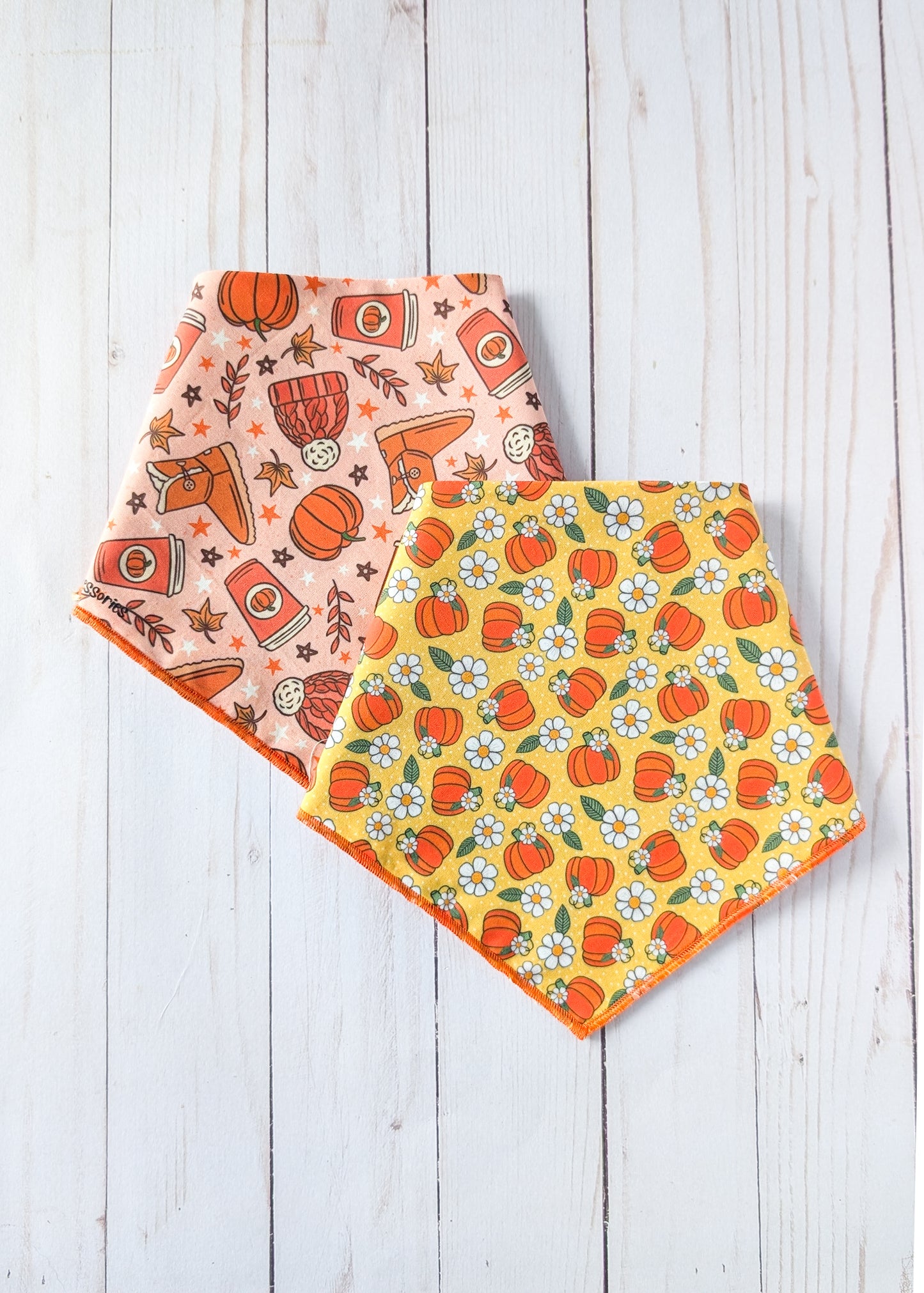 Two triangle bandanas with autumn-themed patterns, one in peach with orange pumpkins and brown boots, and one in yellow with orange pumpkins and white flowers, displayed on a white wooden background.