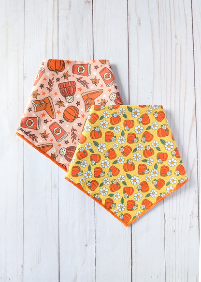 Two triangle bandanas with autumn-themed patterns, one in peach with orange pumpkins and brown boots, and one in yellow with orange pumpkins and white flowers, displayed on a white wooden background.