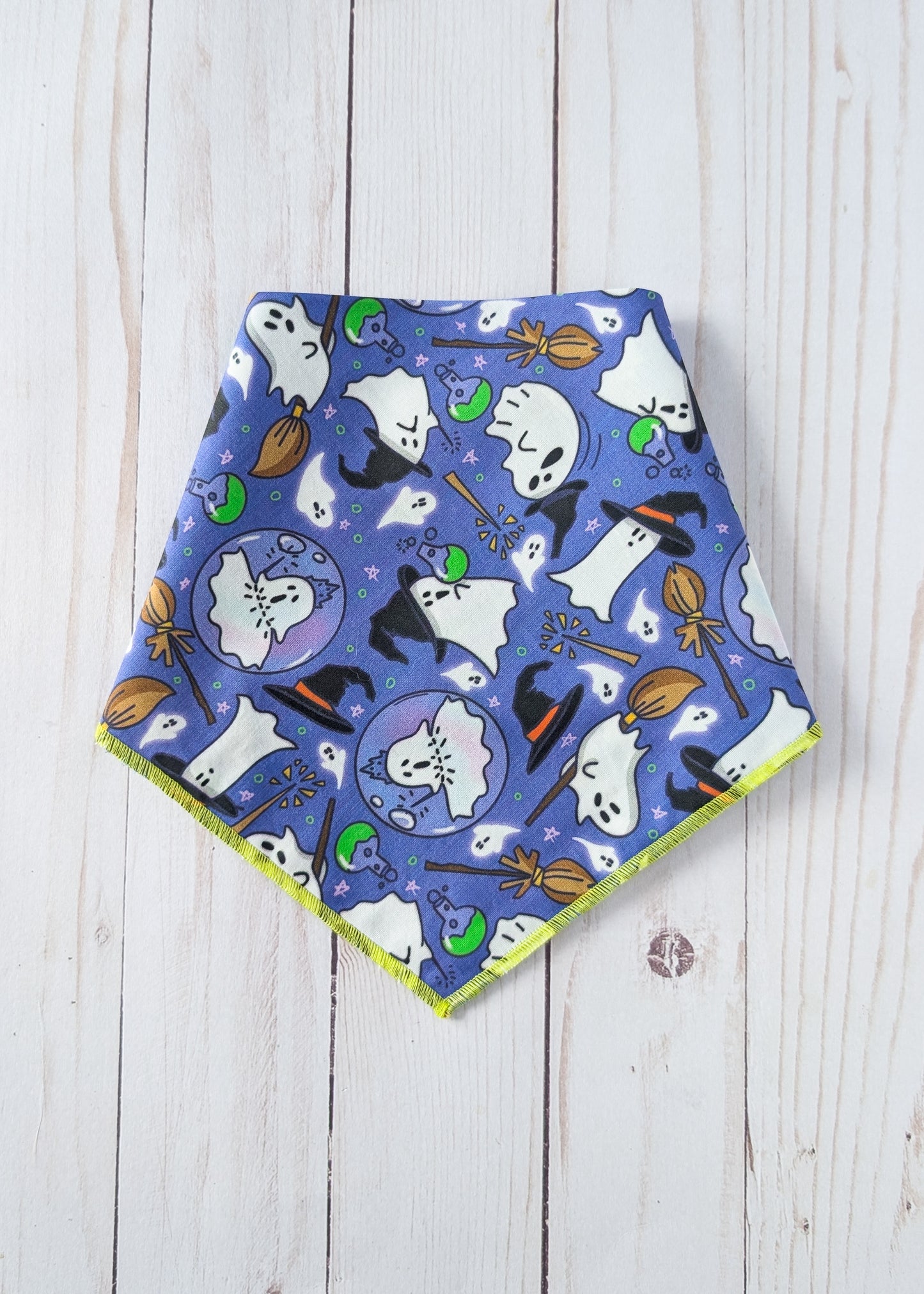 A triangular dog bandana with a violet Halloween pattern featuring ghosts, witches, broomsticks, and pumpkins, displayed flat on a white wooden background.