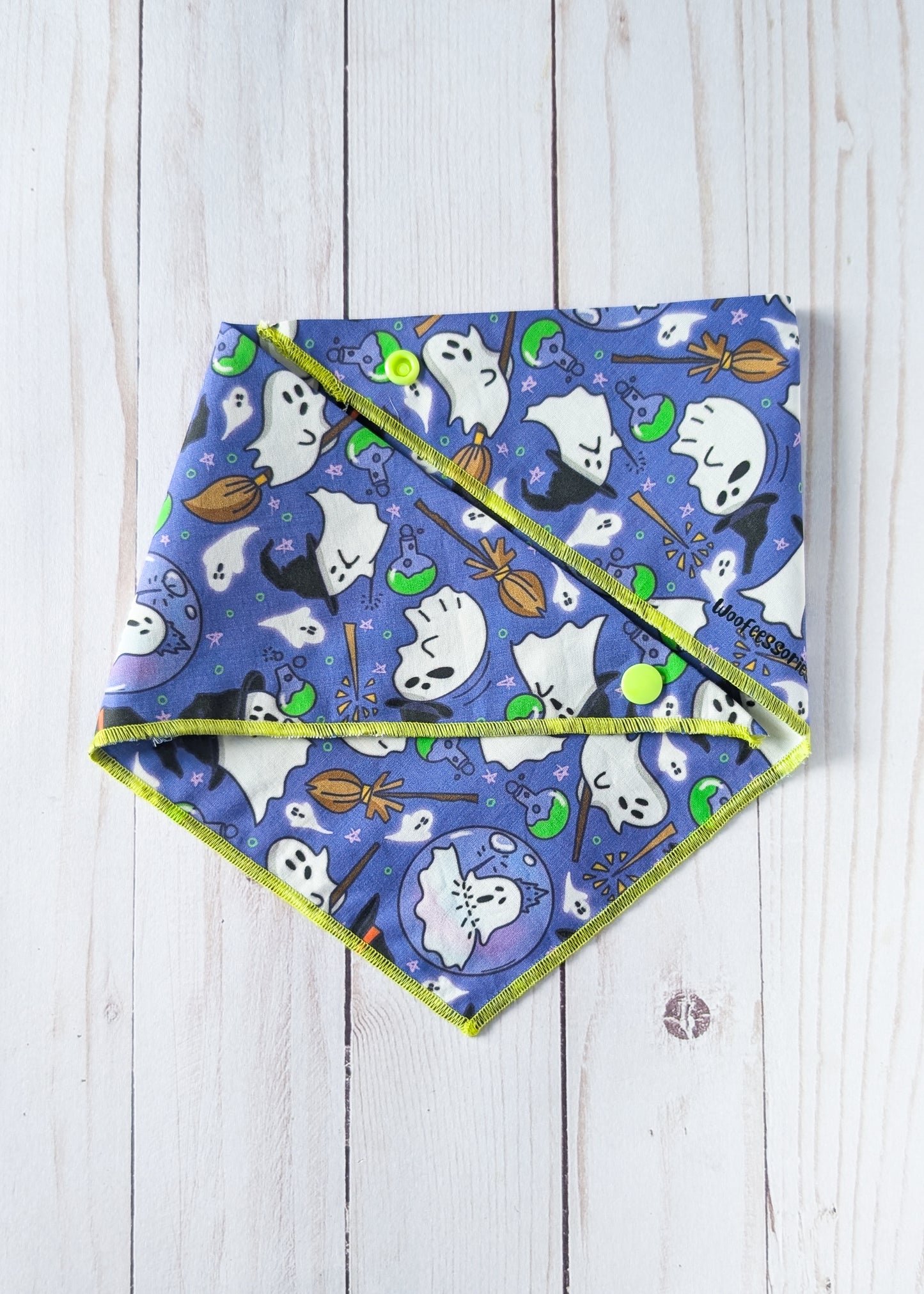 A triangular pet bandana with a blue and purple Halloween print of ghosts, bats, broomsticks, and pumpkins, finished with a lime green trim, laid flat on a white wood background.