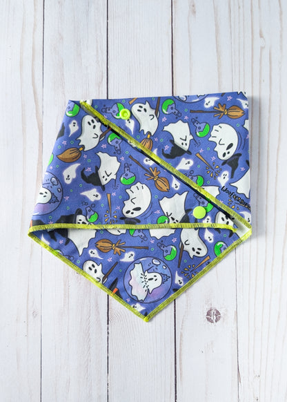 A triangular pet bandana with a blue and purple Halloween print of ghosts, bats, broomsticks, and pumpkins, finished with a lime green trim, laid flat on a white wood background.