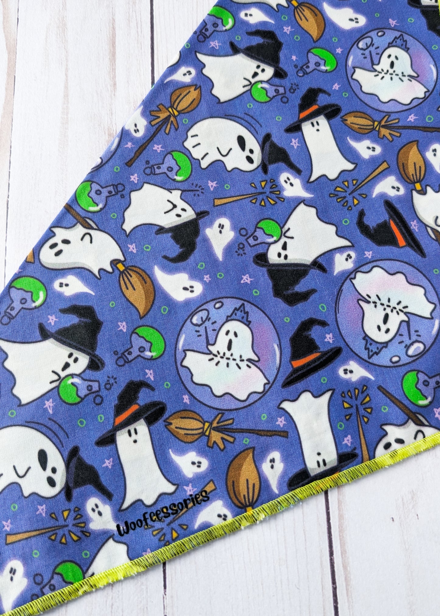 A triangular fabric pet bandana with a purple Halloween print featuring white ghosts, black witch hats, orange brooms, laid flat on a white wood background.