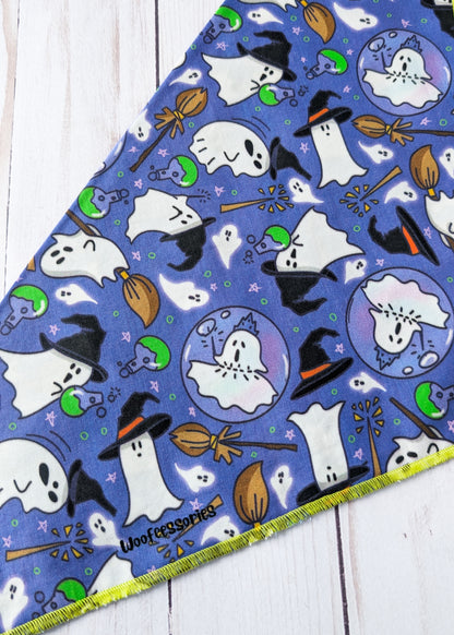 A triangular fabric pet bandana with a purple Halloween print featuring white ghosts, black witch hats, orange brooms, laid flat on a white wood background.