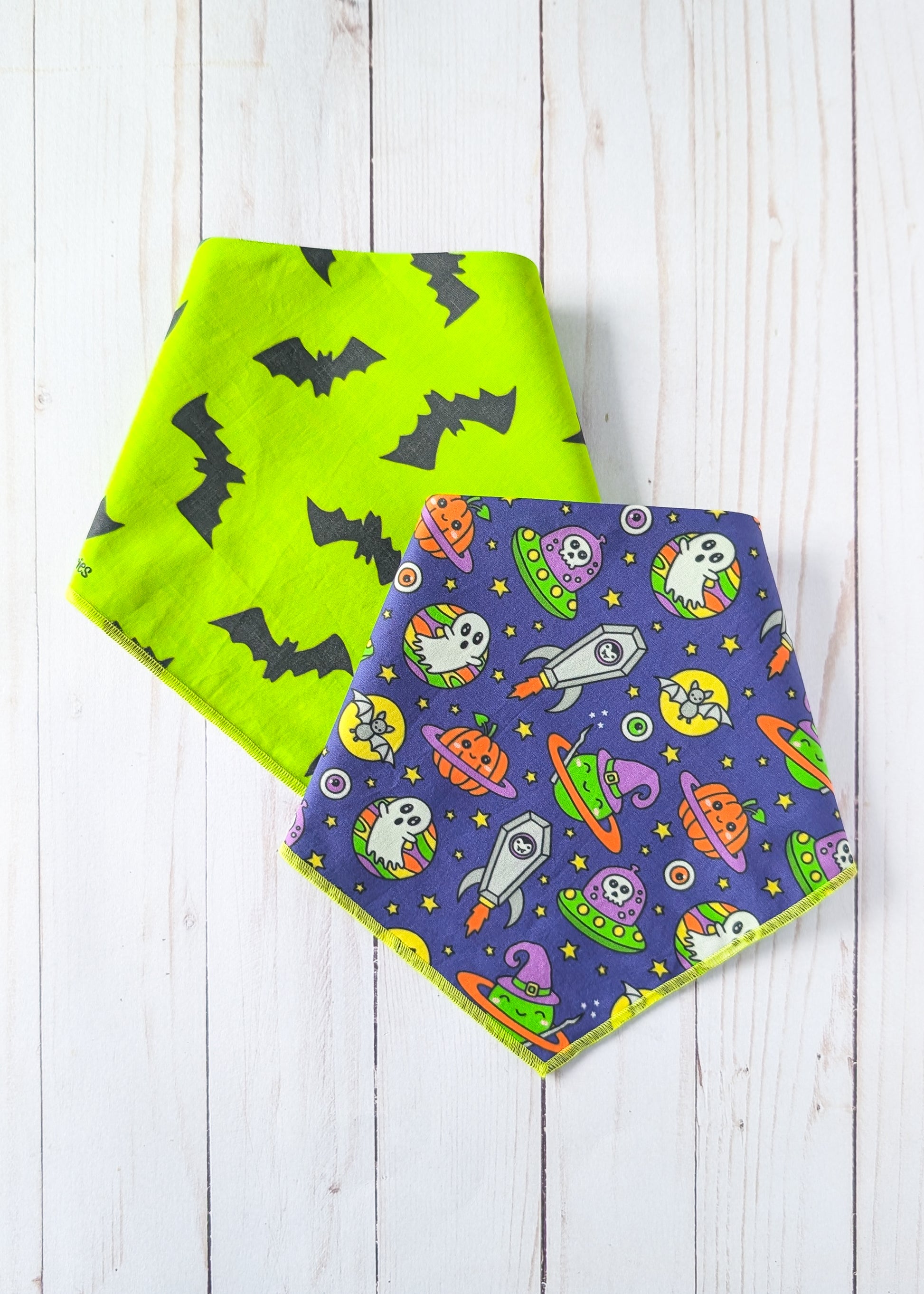 Two triangular bandana bibs with a Halloween theme, one in bright lime green with black bats and the other in dark purple with space and ghost motifs, displayed on a white wood plank background.