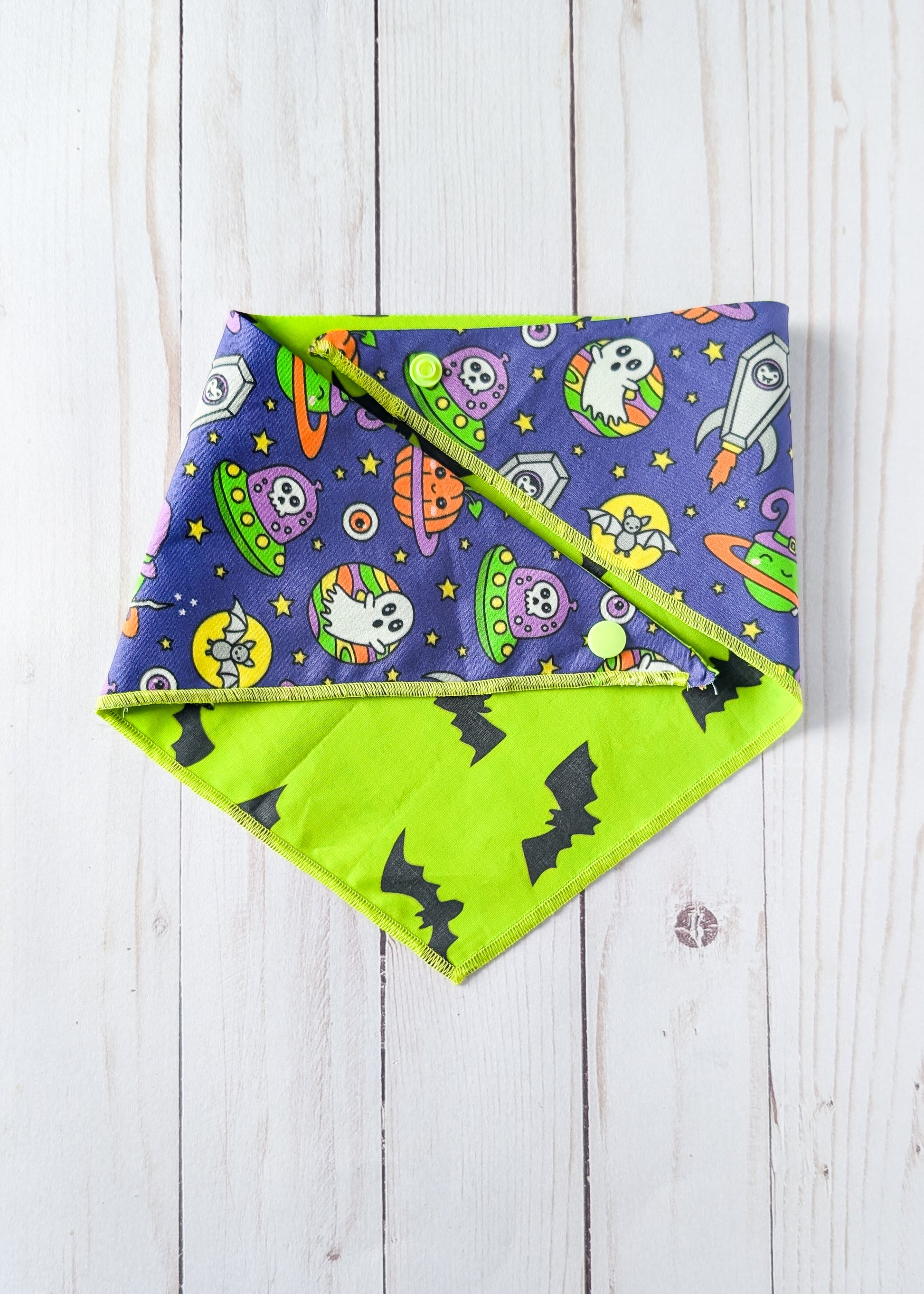 A triangular reversible dog bandana with a Halloween pattern, featuring one side with cartoon ghosts, aliens, and rockets, and a bright lime reverse side with black bat silhouettes, laid flat on a white wood plank background.