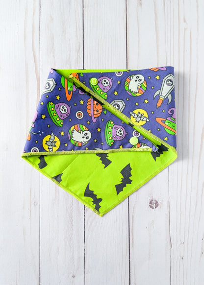 A triangular reversible dog bandana with a Halloween pattern, featuring one side with cartoon ghosts, aliens, and rockets, and a bright lime reverse side with black bat silhouettes, laid flat on a white wood plank background.