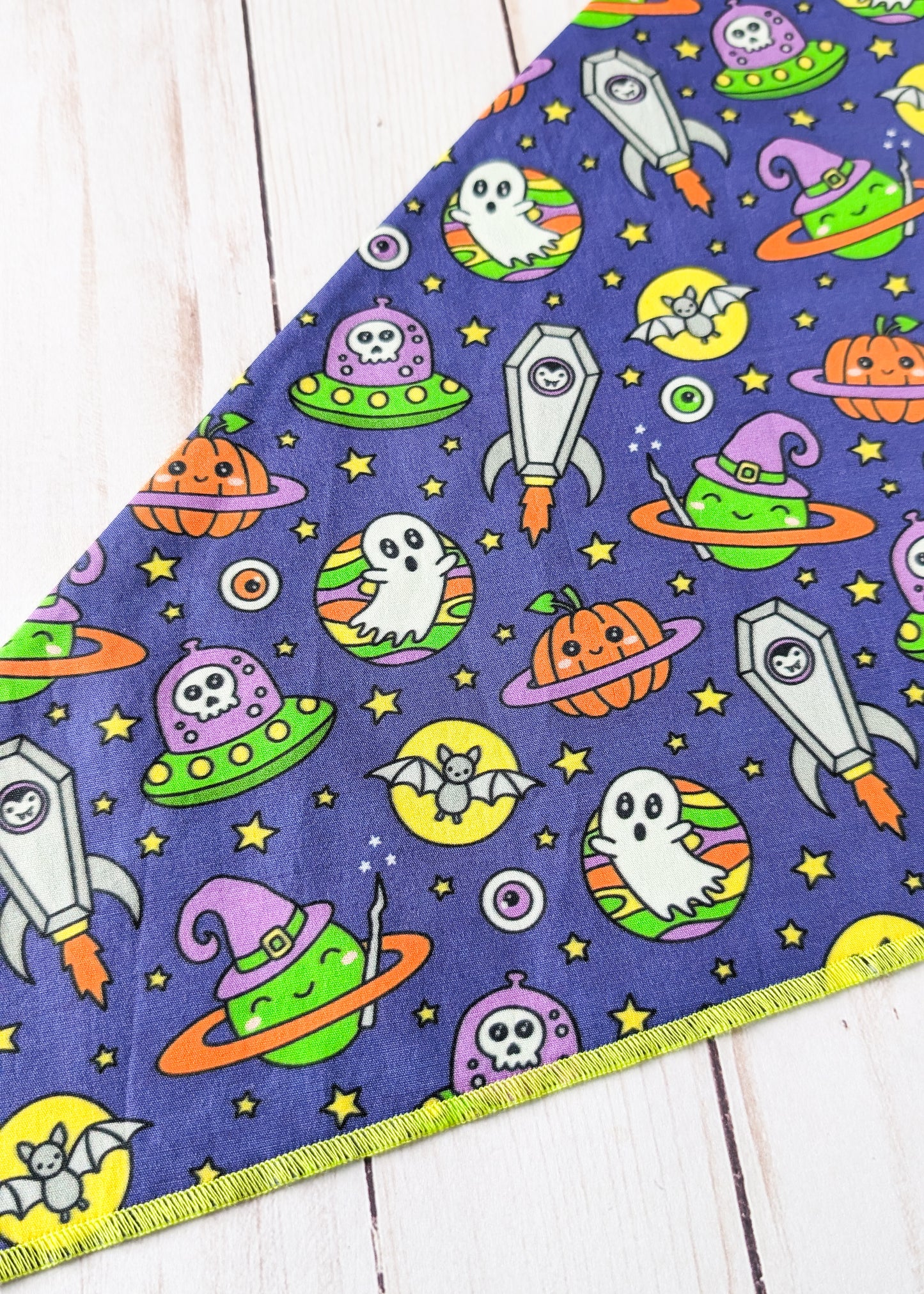 A triangular fabric bandana or pet bandana featuring a colorful Halloween space print on a dark purple background, laid flat on a white wood plank surface.