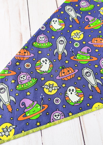 A triangular fabric bandana or pet bandana featuring a colorful Halloween space print on a dark purple background, laid flat on a white wood plank surface.