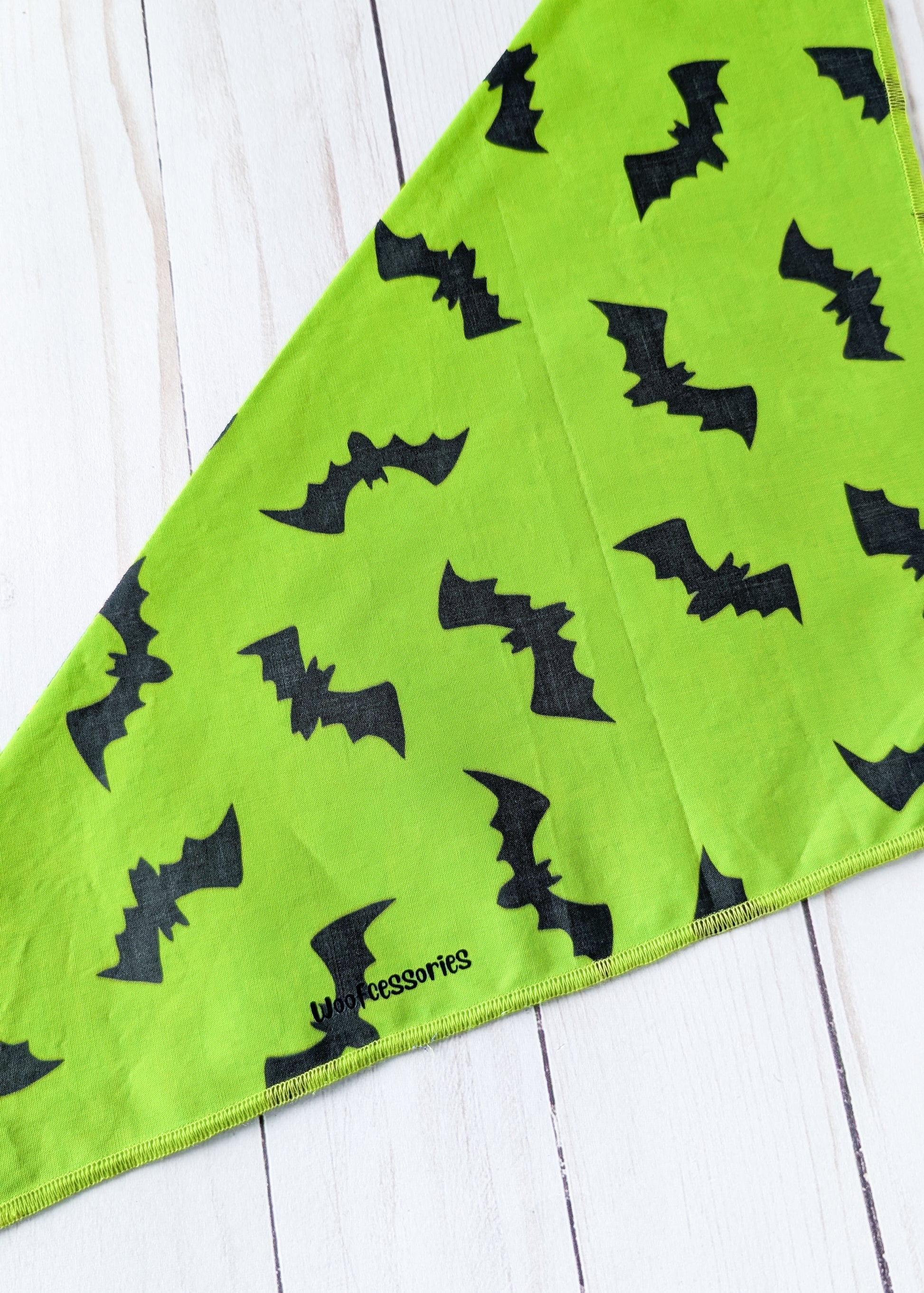 A lime green triangular bandana featuring a black bat pattern, laid flat on a white wood plank surface