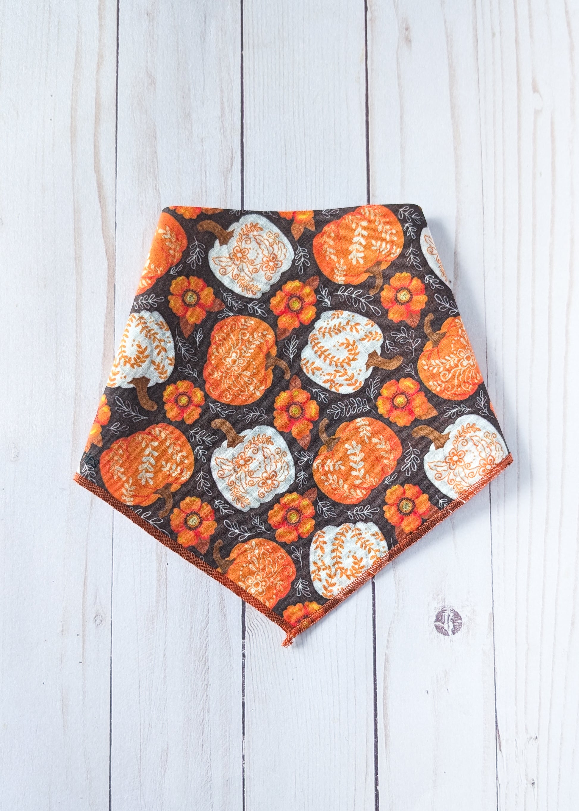 A triangular pet bandana with an autumn pattern of orange and white pumpkins and flowers on a dark background, laid flat on a white wood plank surface.