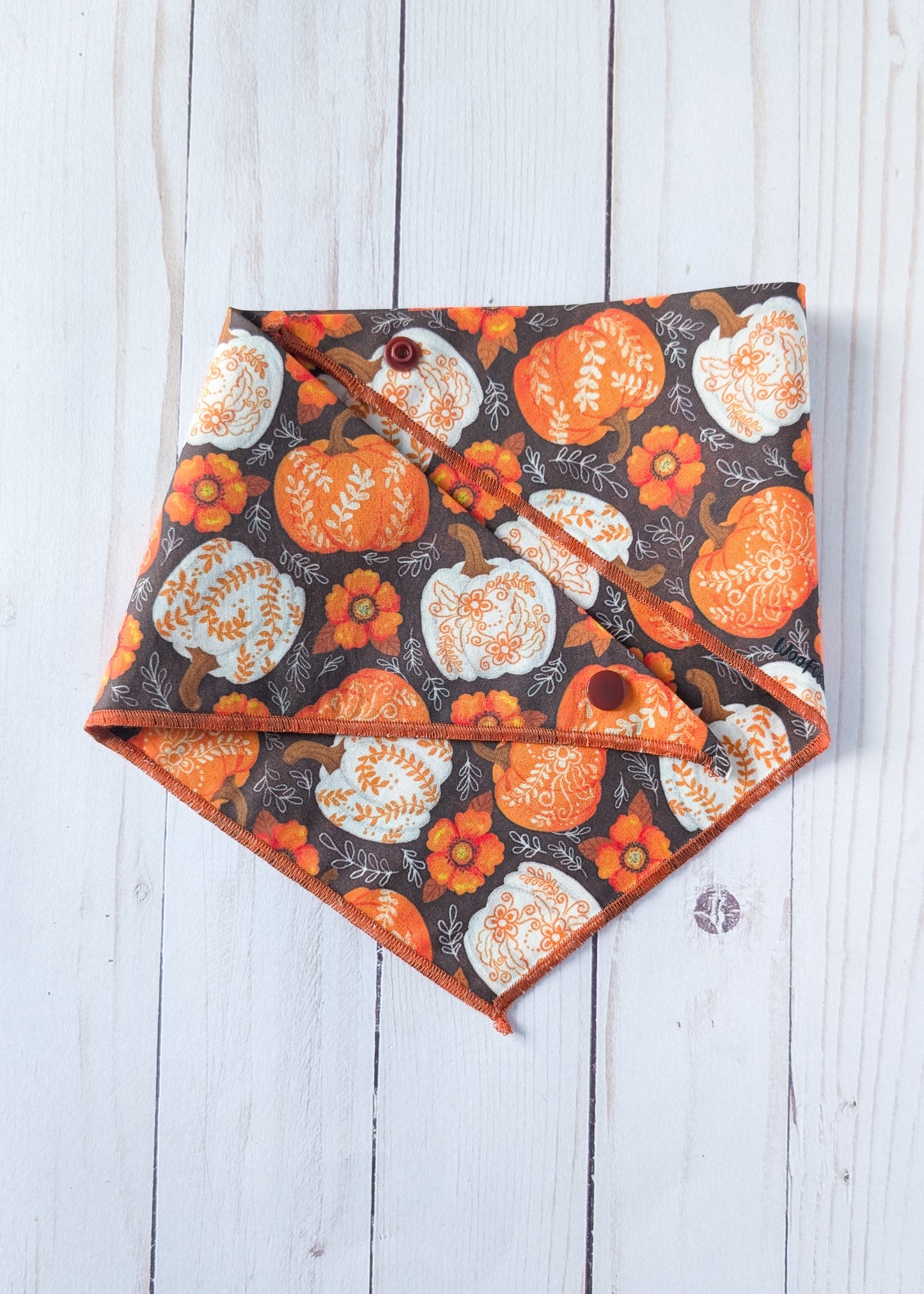 A triangular pet bandana with an autumn pattern of orange and white pumpkins and flowers on a dark background, laid flat on a white wood plank surface.