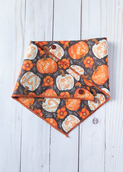 A triangular pet bandana with an autumn pattern of orange and white pumpkins and flowers on a dark background, laid flat on a white wood plank surface.