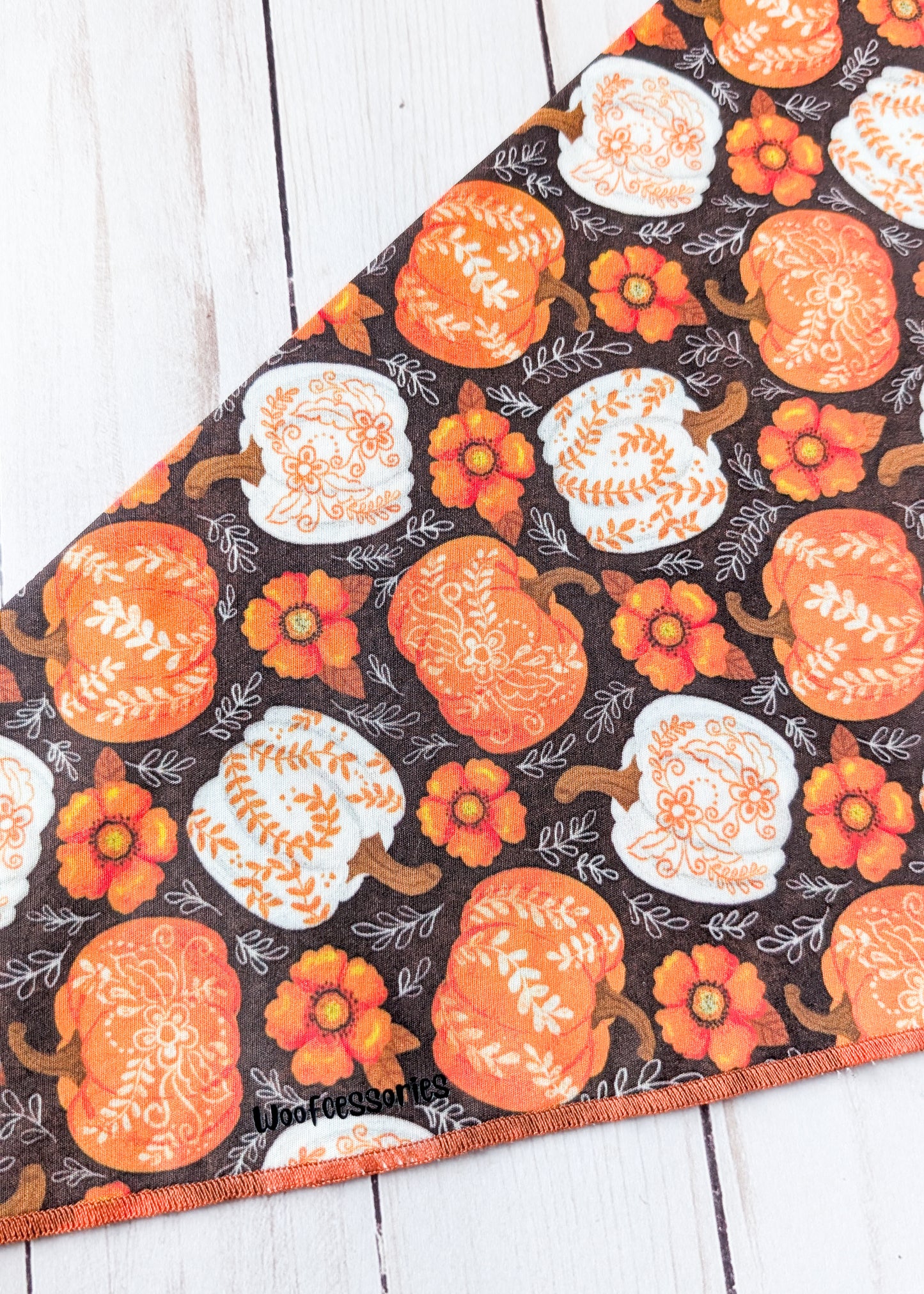 A close up of a triangular pet bandana with an autumn pattern of orange and white pumpkins and flowers on a dark background, laid flat on a white wood plank surface.