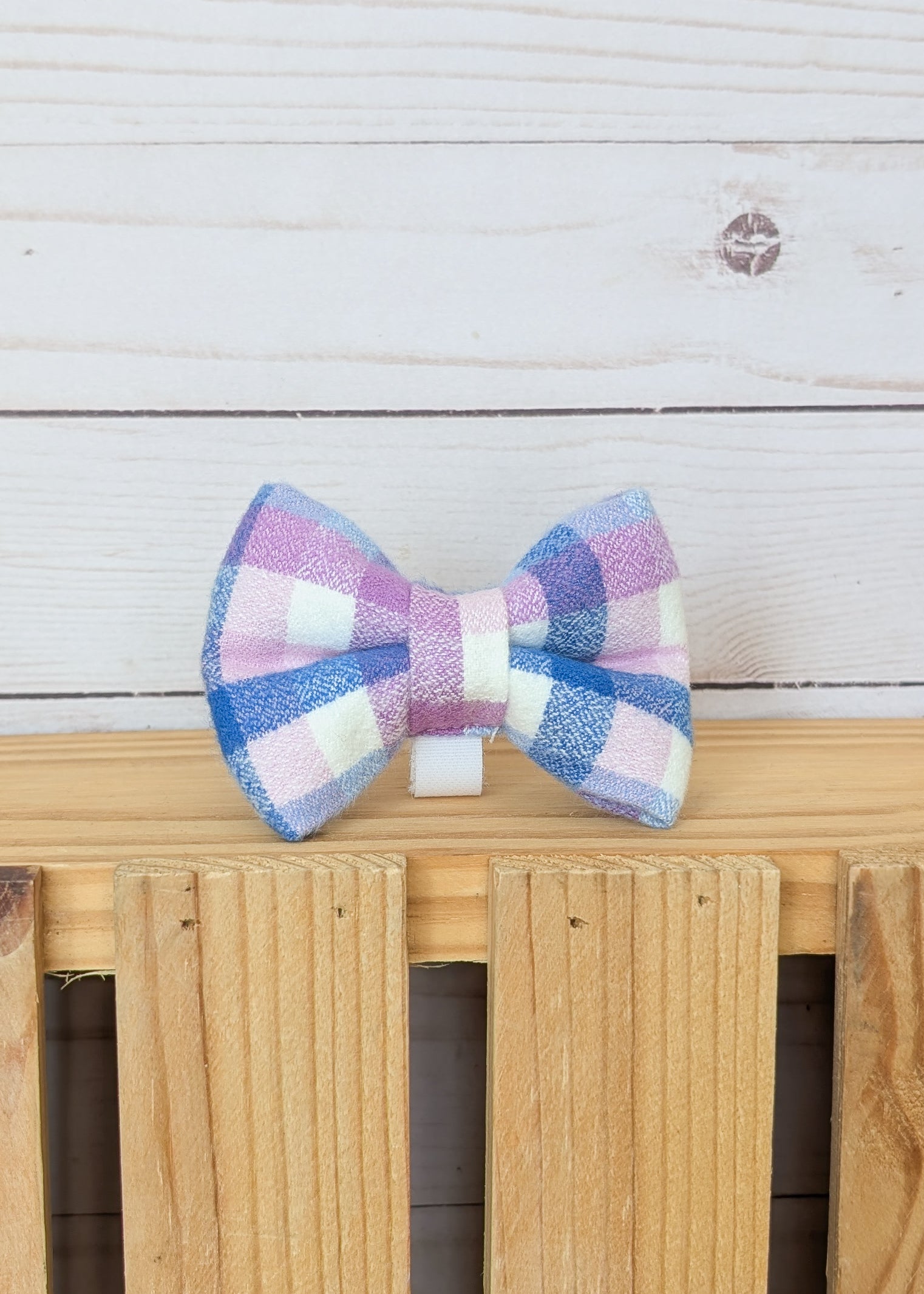 A small bow tie with a pastel pink, purple, blue, and white gingham pattern, displayed on a light wood surface against a white wood plank background.