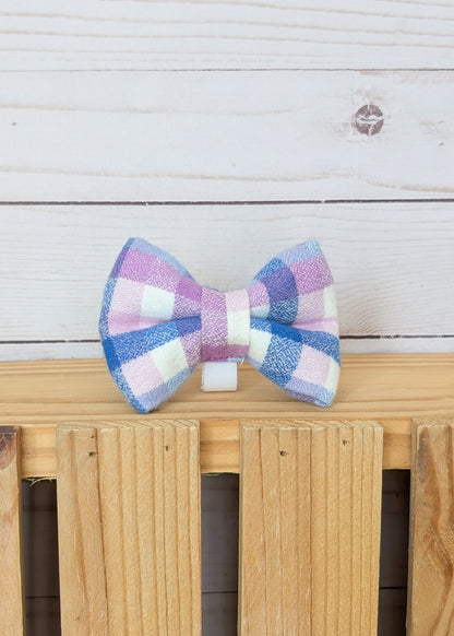 A small bow tie with a pastel pink, purple, blue, and white gingham pattern, displayed on a light wood surface against a white wood plank background.
