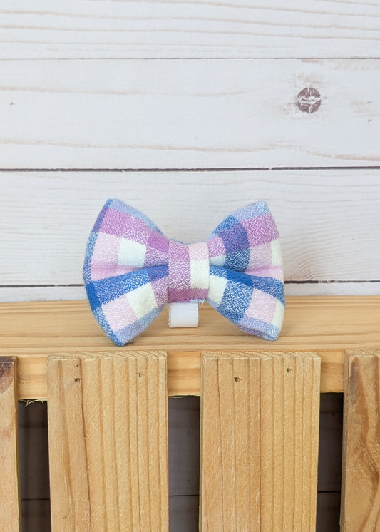 A small bow tie with a pastel pink, purple, blue, and white gingham pattern, displayed on a light wood surface against a white wood plank background.