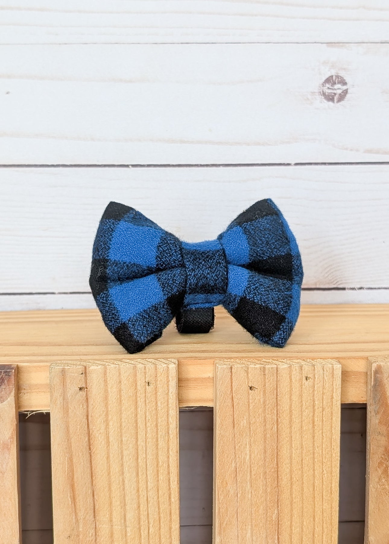 A blue and black buffalo plaid bow tie displayed on a light wooden slatted surface against a white wood plank background.