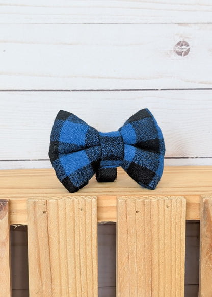 A blue and black buffalo plaid bow tie displayed on a light wooden slatted surface against a white wood plank background.