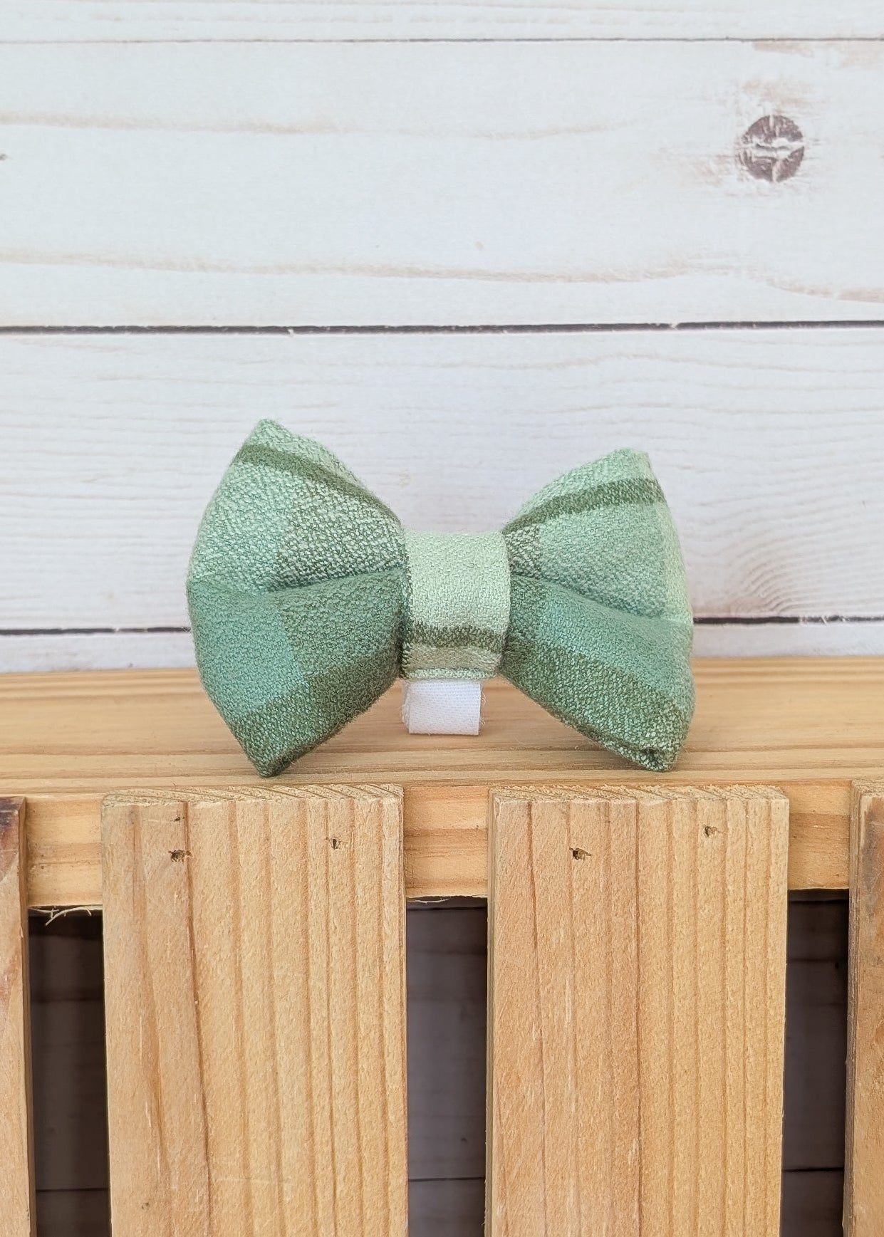 A light green plaid flannel fabric bow tie resting on a wooden slatted surface against a white wood plank background.