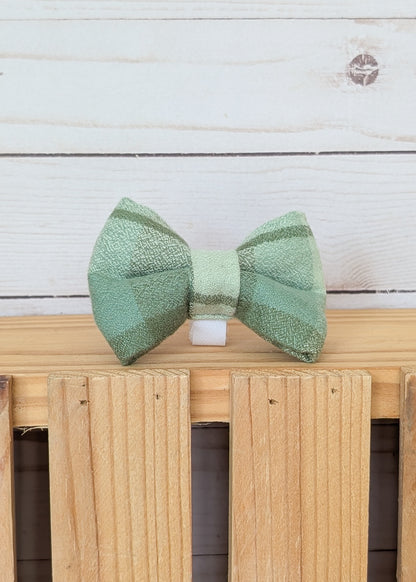 A light green plaid flannel fabric bow tie resting on a wooden slatted surface against a white wood plank background.